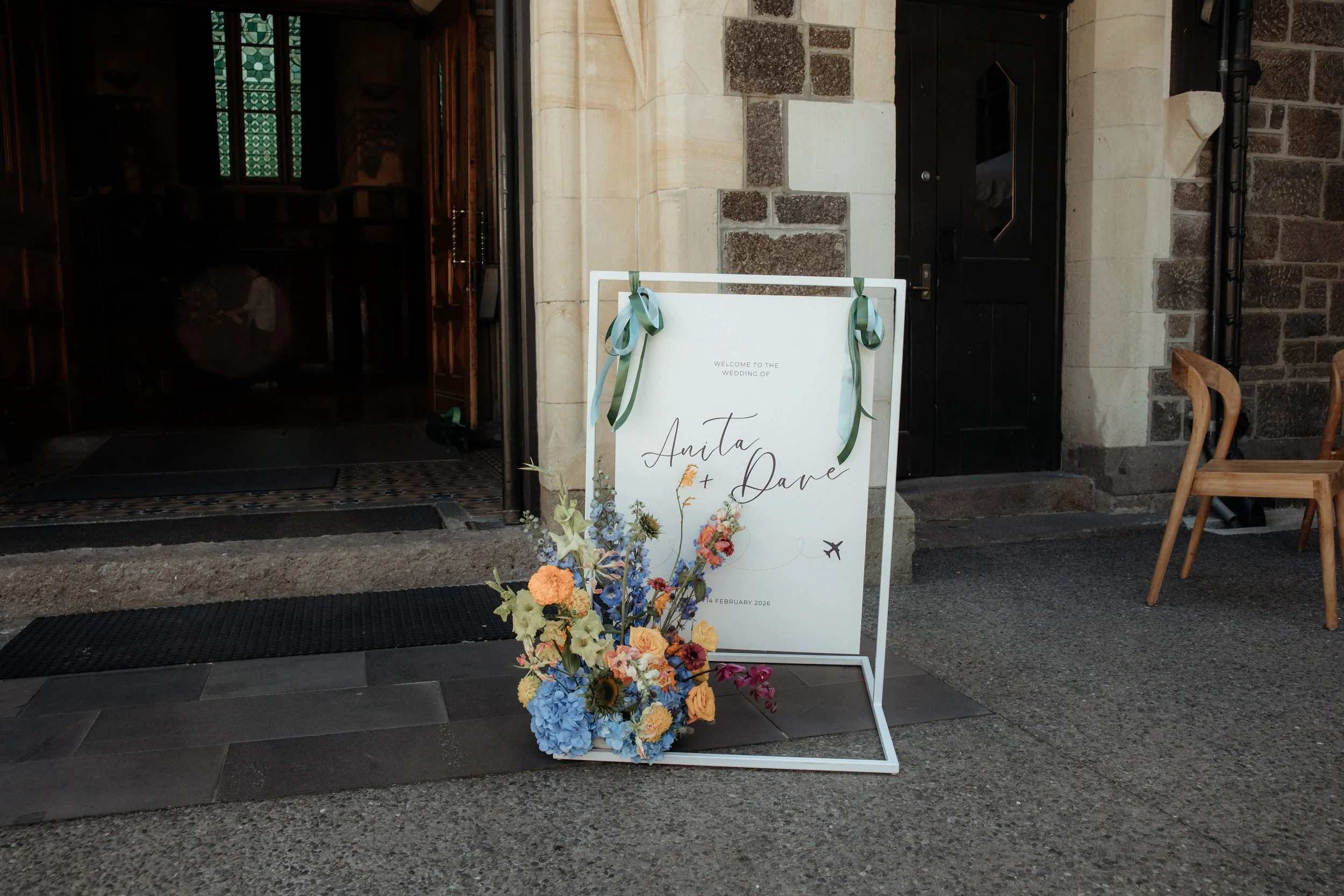 Wedding welcome sign with floral arrangement in front, outside of a building with stone walls and black door.