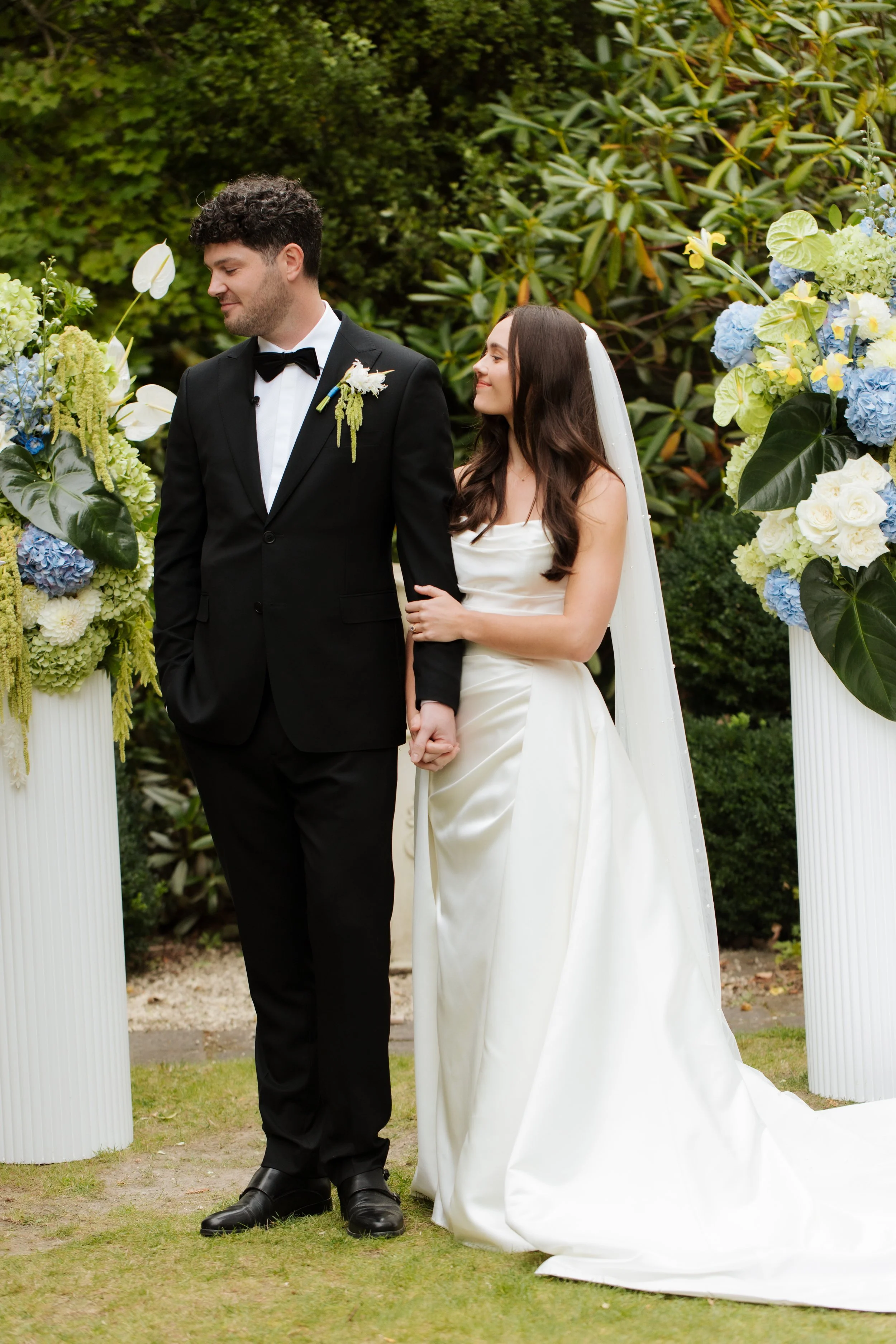A newlywed couple standing outdoors, holding hands, with a floral arch behind them. The groom in a black tuxedo and the bride in a white wedding dress with a veil, surrounded by green foliage.