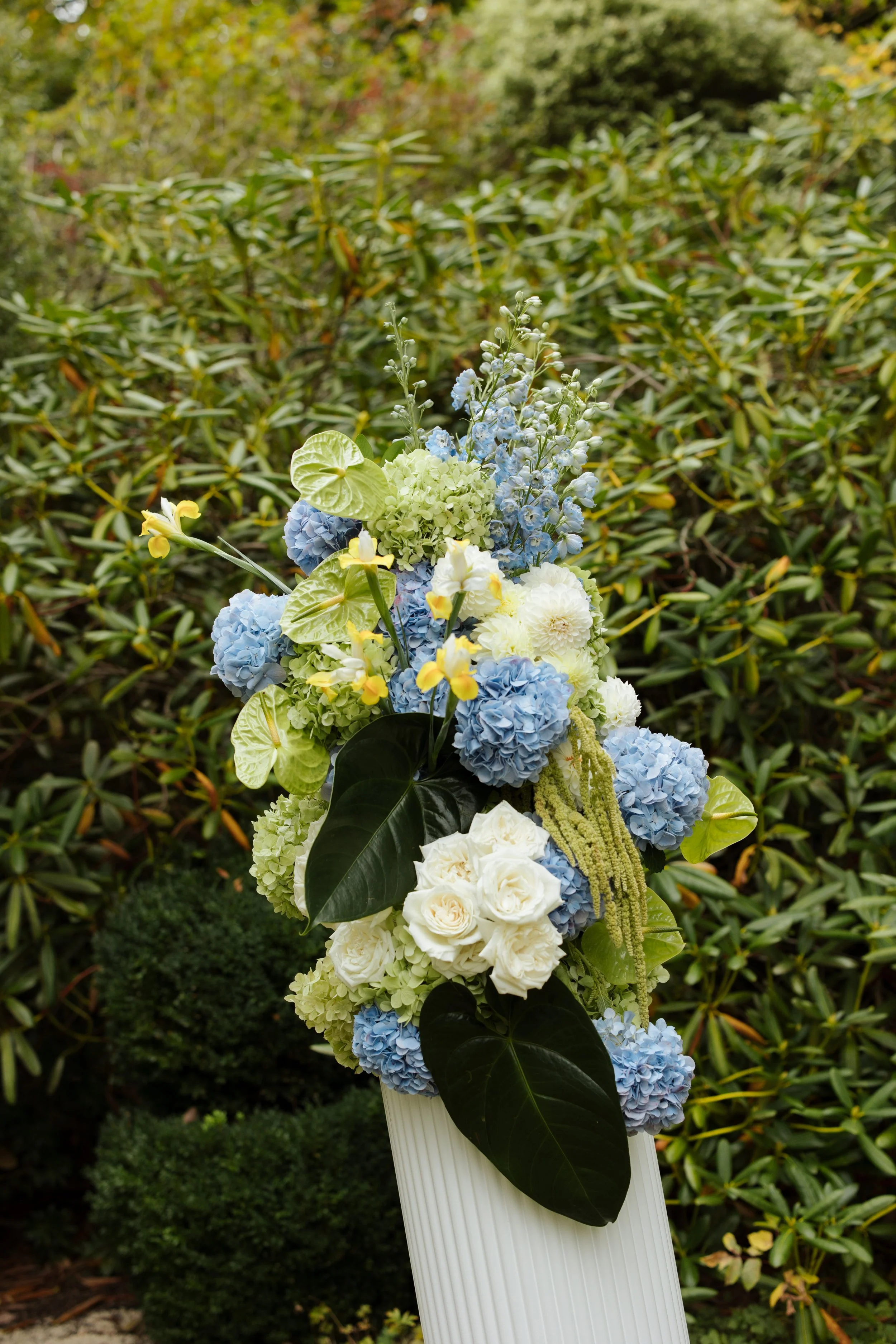 A floral arrangement of white, blue, and yellow flowers with green leaves, placed on a white stand outdoors, with green shrubbery in the background.