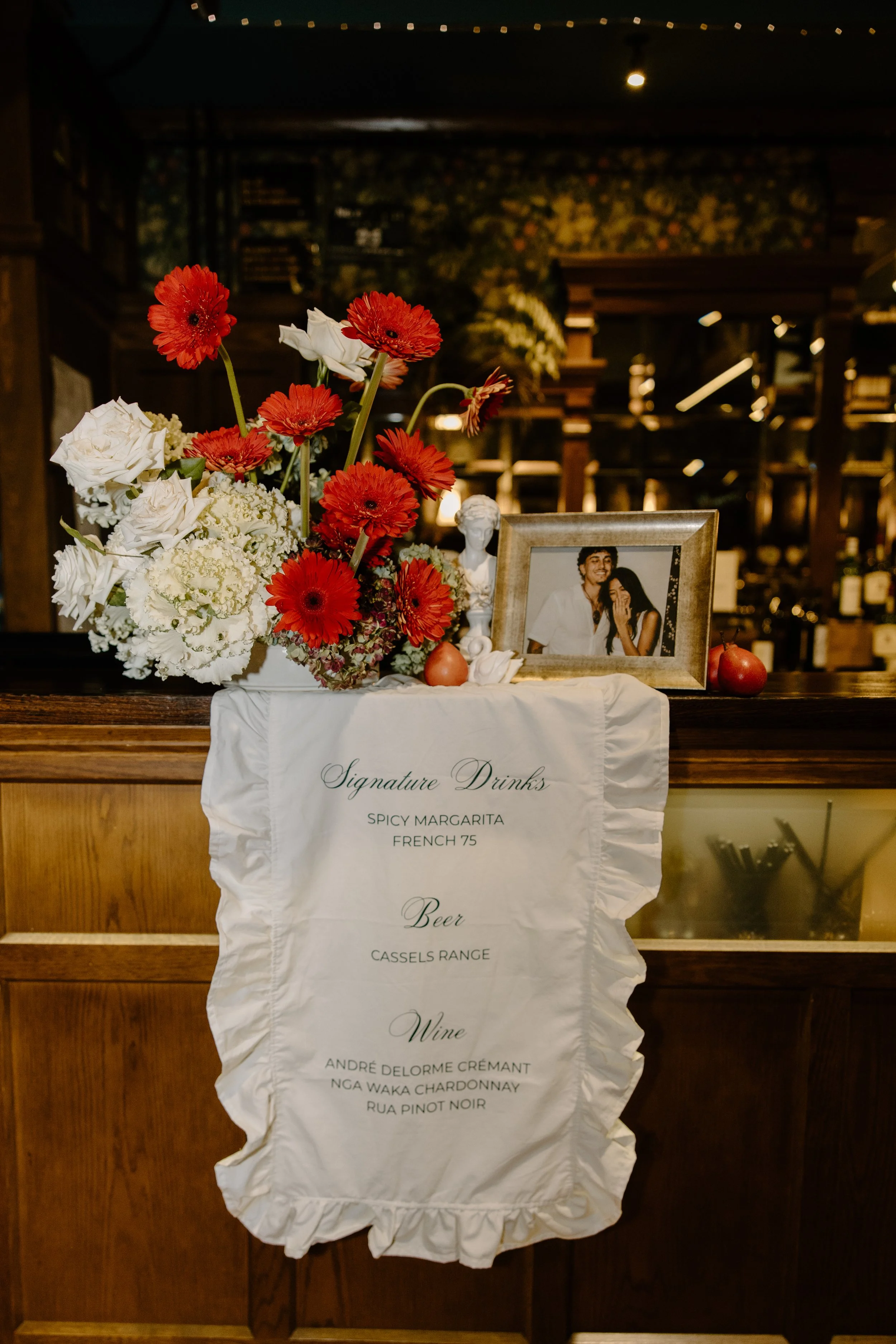 A floral arrangement with red and white flowers, a framed photograph of a couple, and a menu listing signature drinks, beer, and wine on a white cloth draped over a wooden surface in a dimly lit setting.