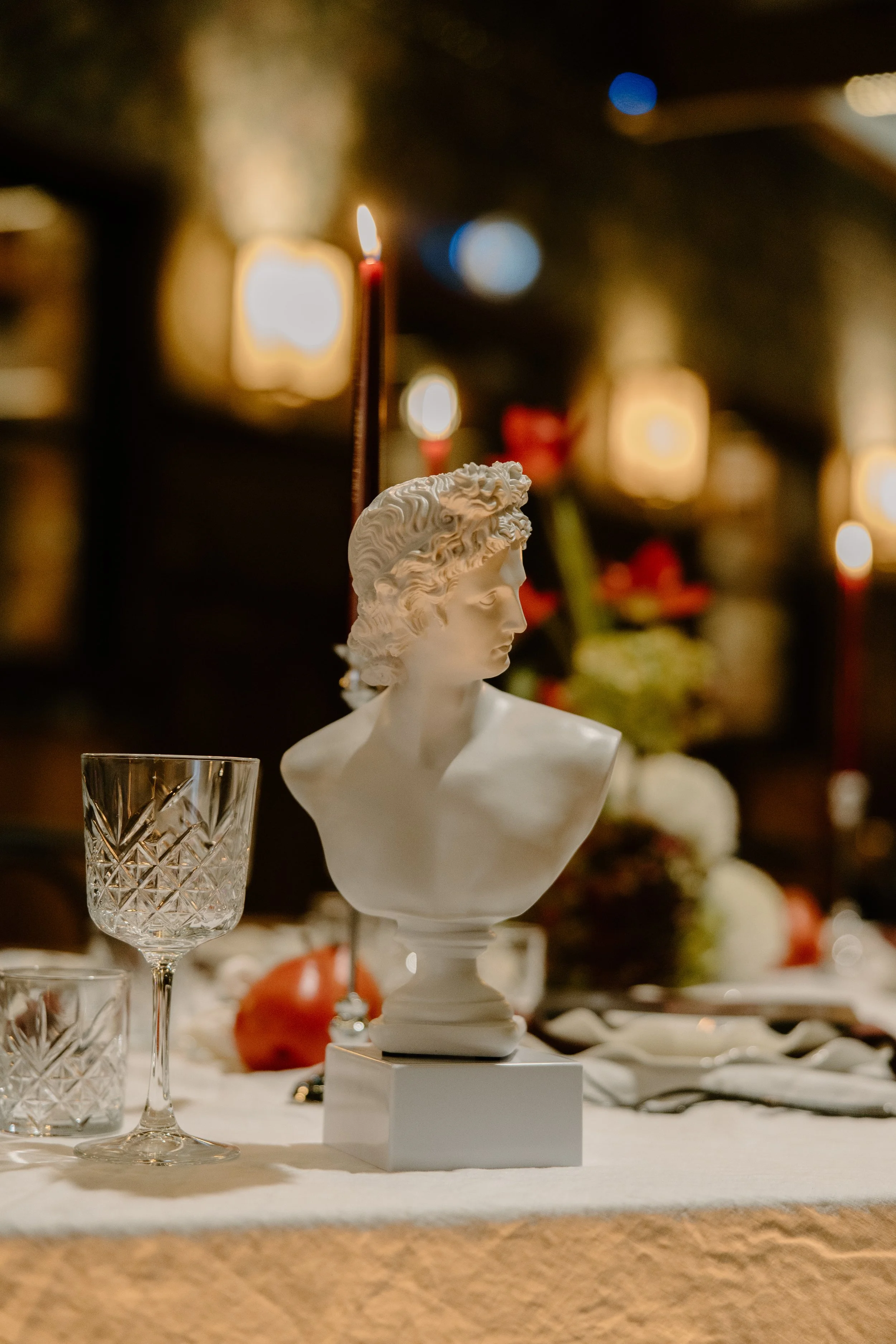 A white bust sculpture of a woman's head and shoulders on a white pedestal, surrounded by crystal glasses and a table setting, with blurred warm lighting and decorative objects in the background.
