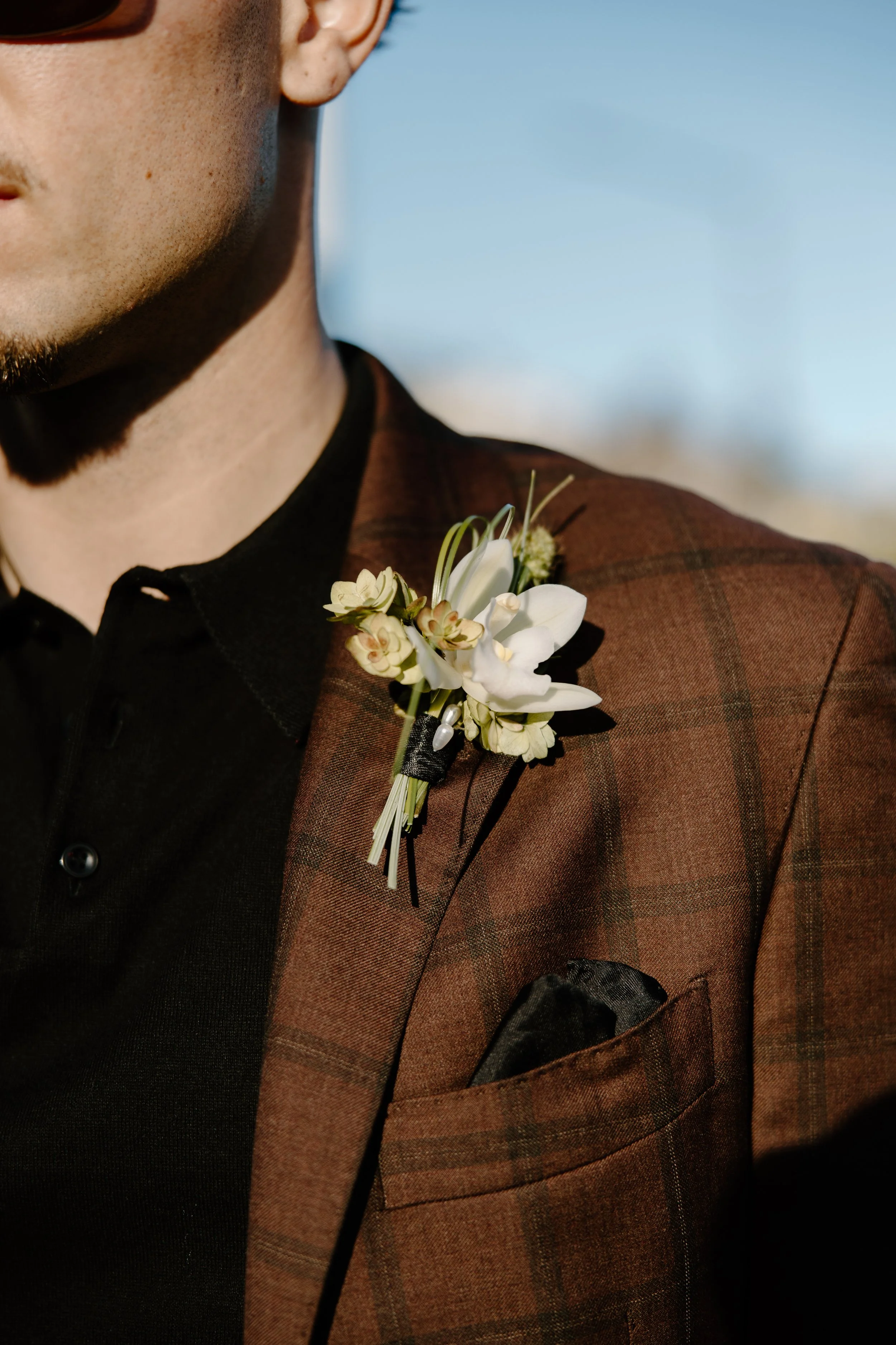 Close-up of a man wearing a brown suit jacket with a floral boutonniere and a black shirt.