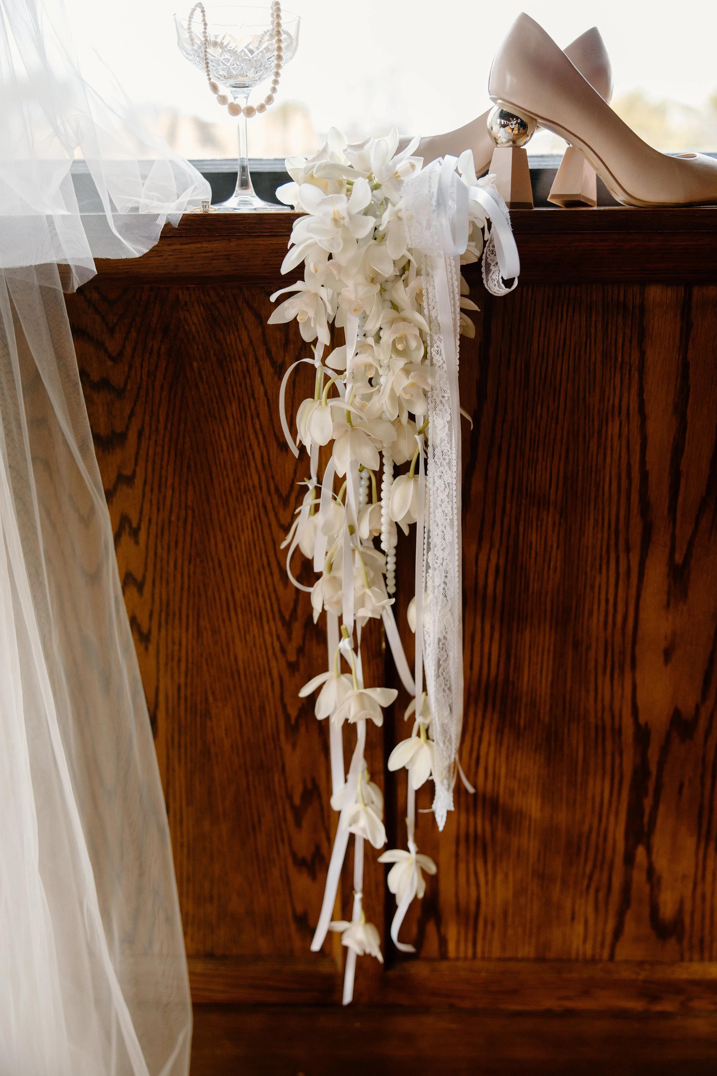 White wedding shoes with decorative heels, a cluster of white orchids, and jewelry on a wooden surface near a window.