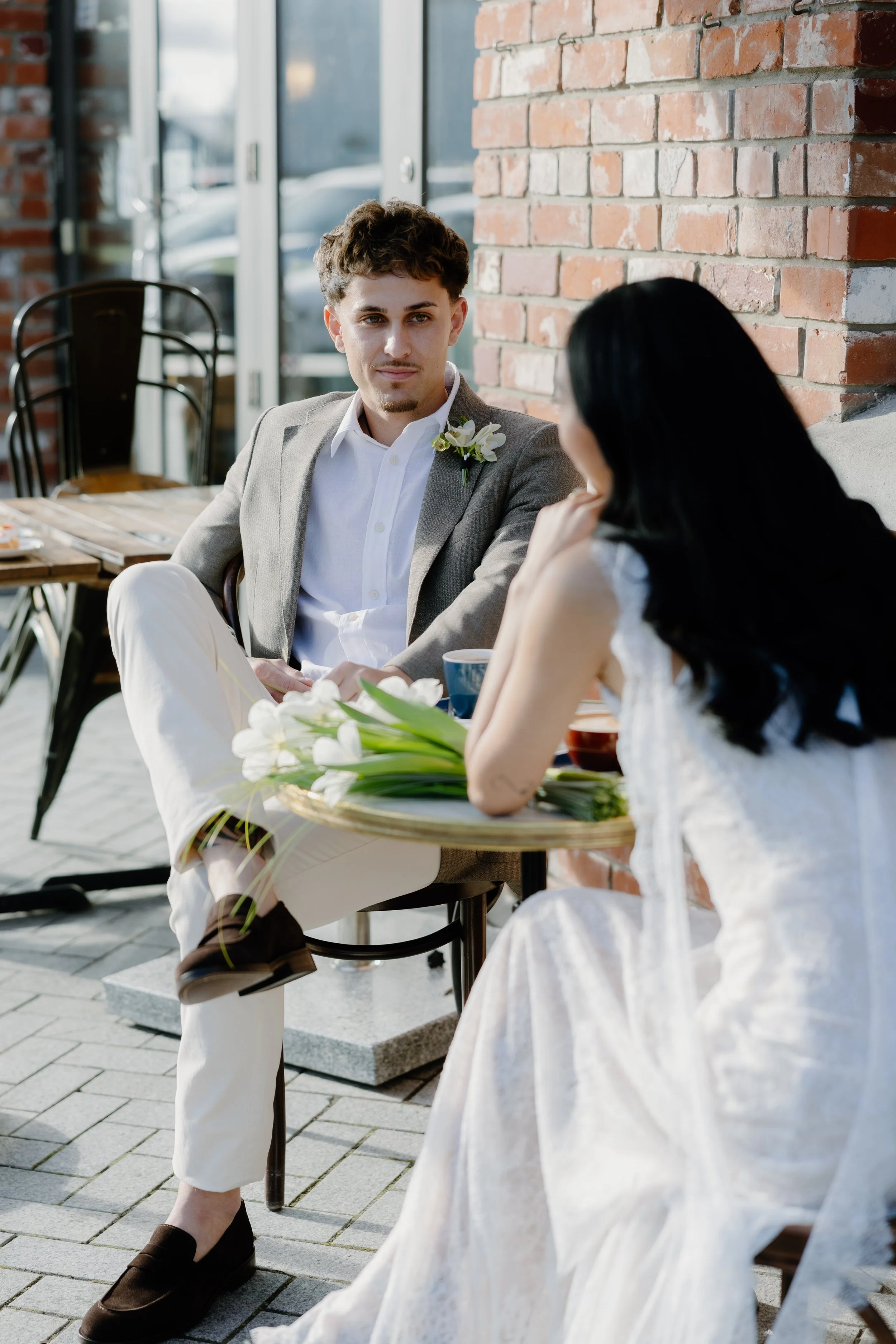 A man in a gray blazer with a boutonniere, white shirt, and beige pants sitting outside at a table, engaged in conversation with a woman in a white lace dress. The scene is set against a brick wall with large windows.