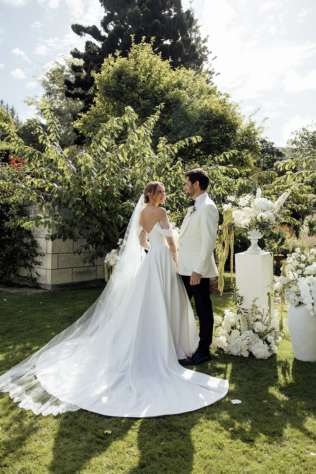 A bride and groom standing outdoors in a garden, facing each other with greenery and white floral arrangements around them, on a sunny day.