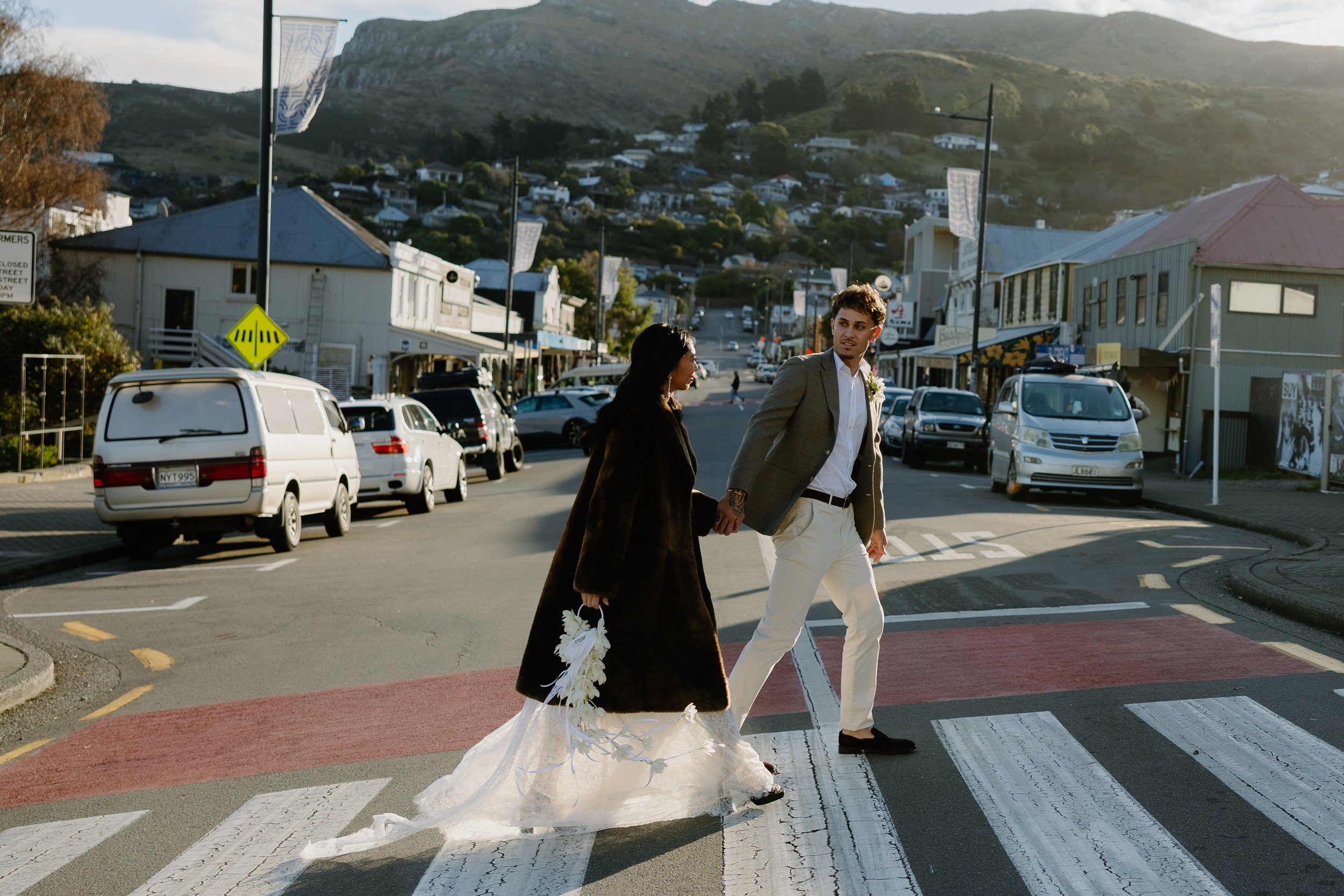 A bride and groom crossing a street at an intersection during what appears to be a wedding celebration, with the bride wearing a wedding dress and holding flowers, and the groom dressed in a suit. The background shows parked cars, buildings, and a hi