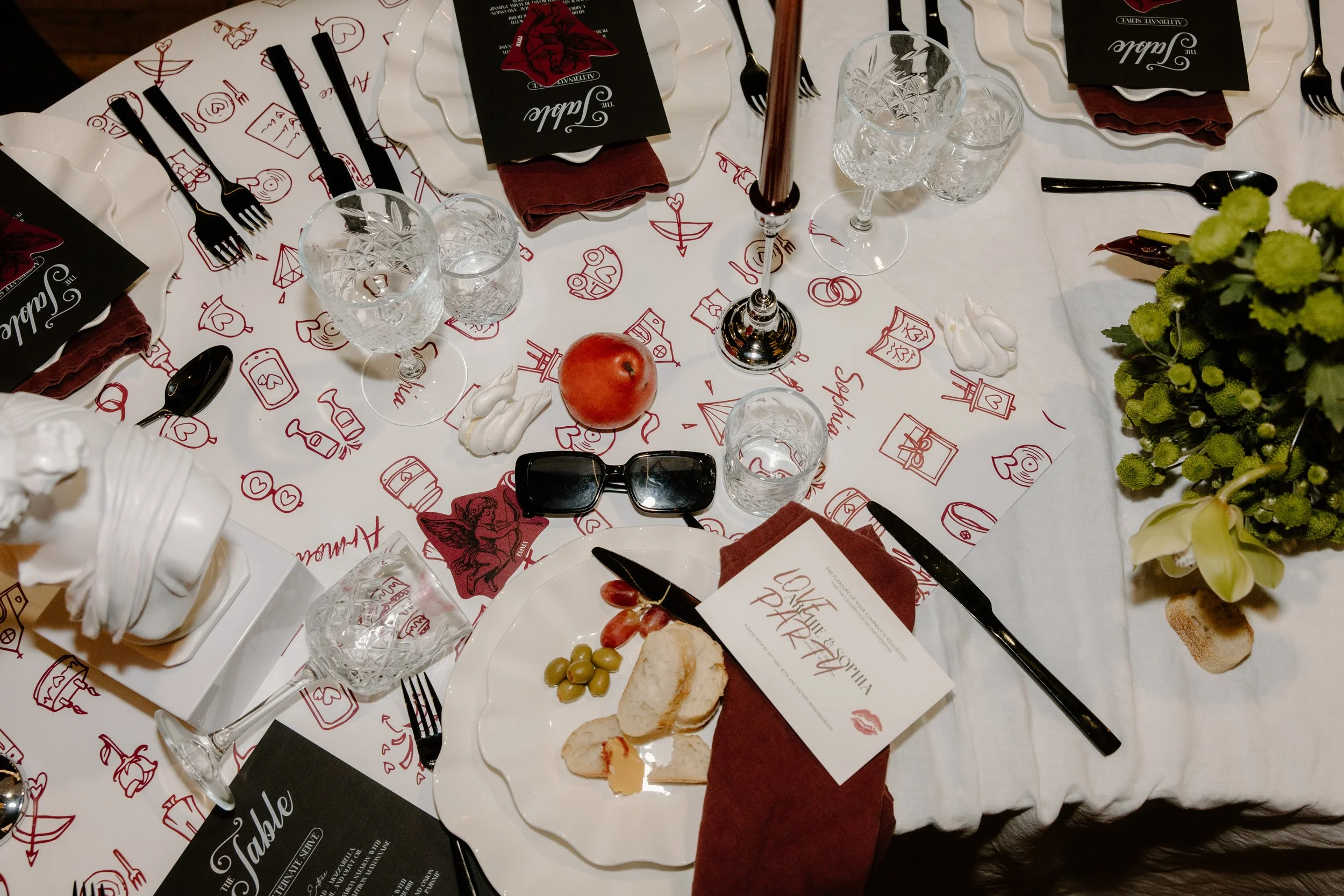 A formal dining table set for a meal with plates, glasses, cutlery, napkins, and a menu. The table has a white tablecloth with red illustrations and a centerpiece with flowers. There are wine glasses, water glasses, a red apple, grapes, and bread on 