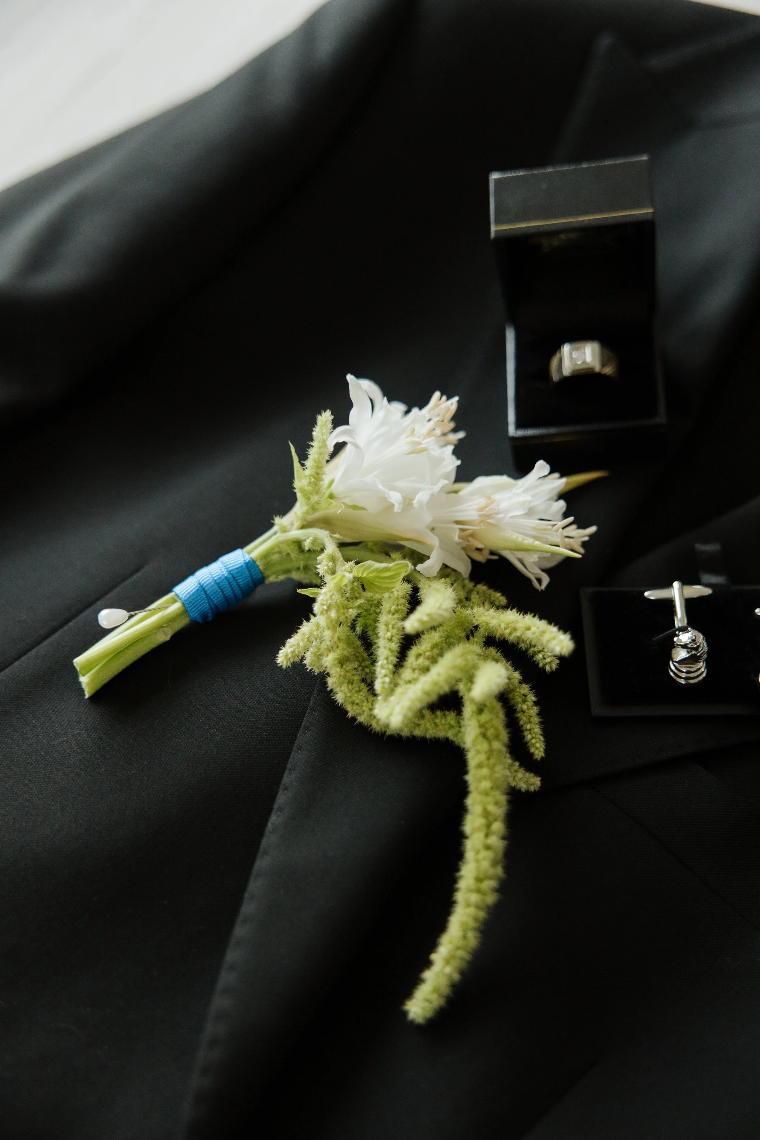 Wedding boutonniere with white flowers, green accents, and blue ribbon, placed next to a small jewelry box with a ring inside, on black fabric background.