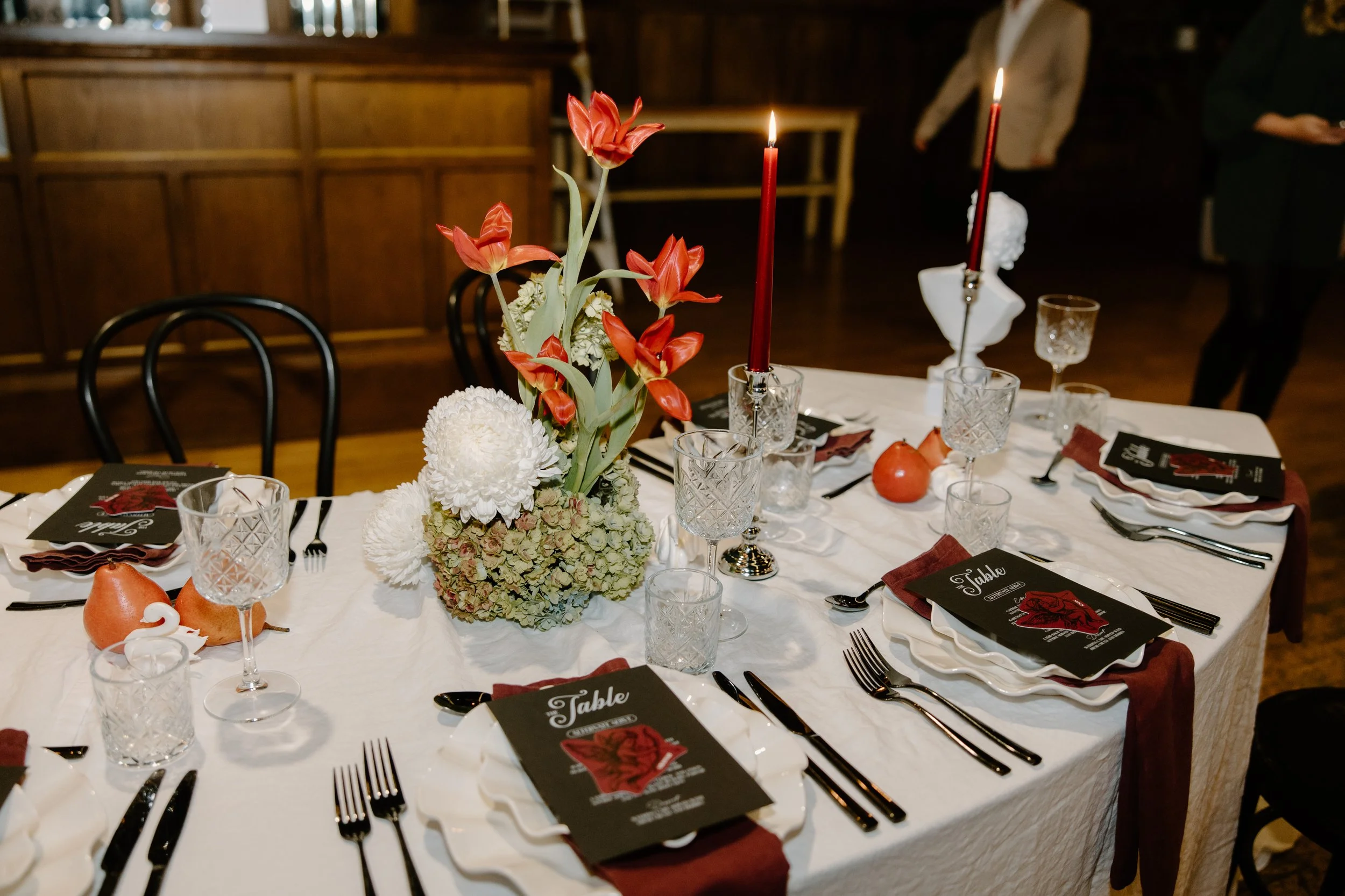 A dining table decorated with a flower arrangement featuring red tulips, white chrysanthemums, and other flowers, along with candles, glassware, and black menus with a red graphic.
