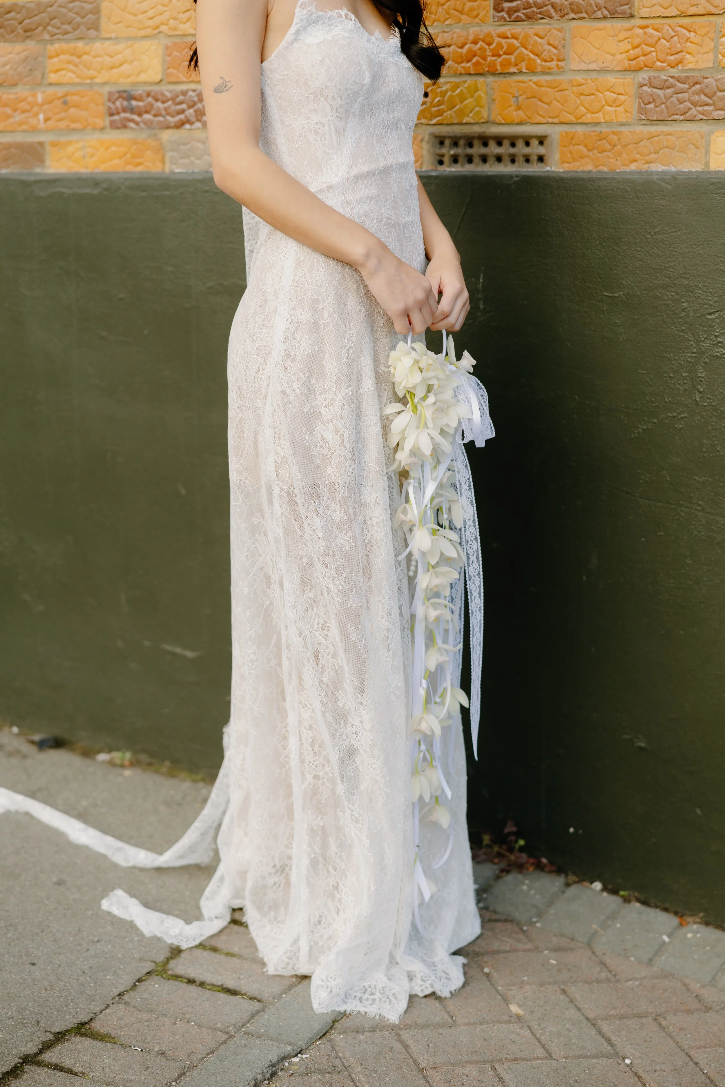 A woman in a white lace dress holding a bouquet of white flowers, standing on a sidewalk against a brick and green wall background.