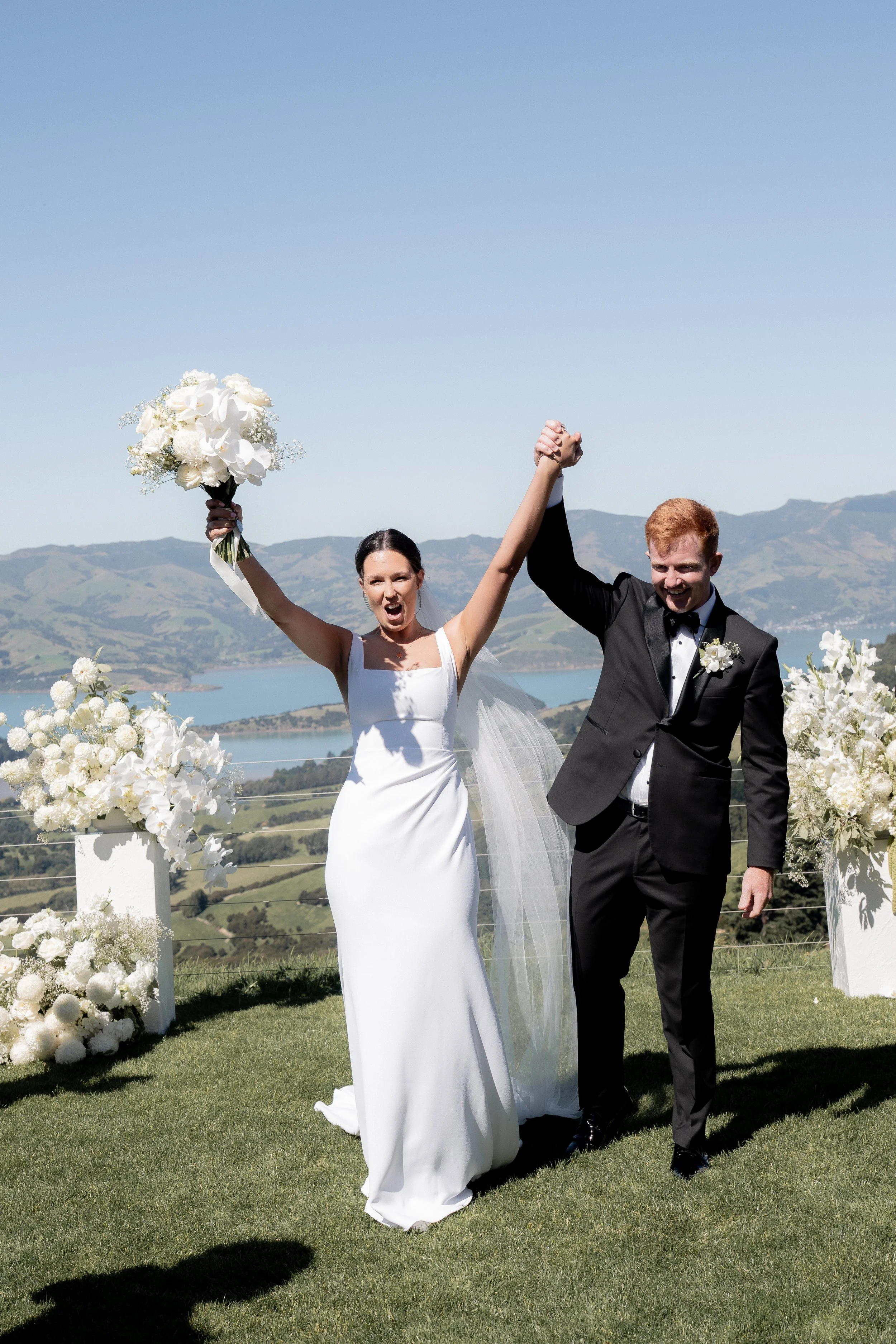A bride and groom celebrating outdoors on their wedding day, holding hands and raising them in victory, with a scenic mountainous landscape and lake in the background, decorated with white flowers.