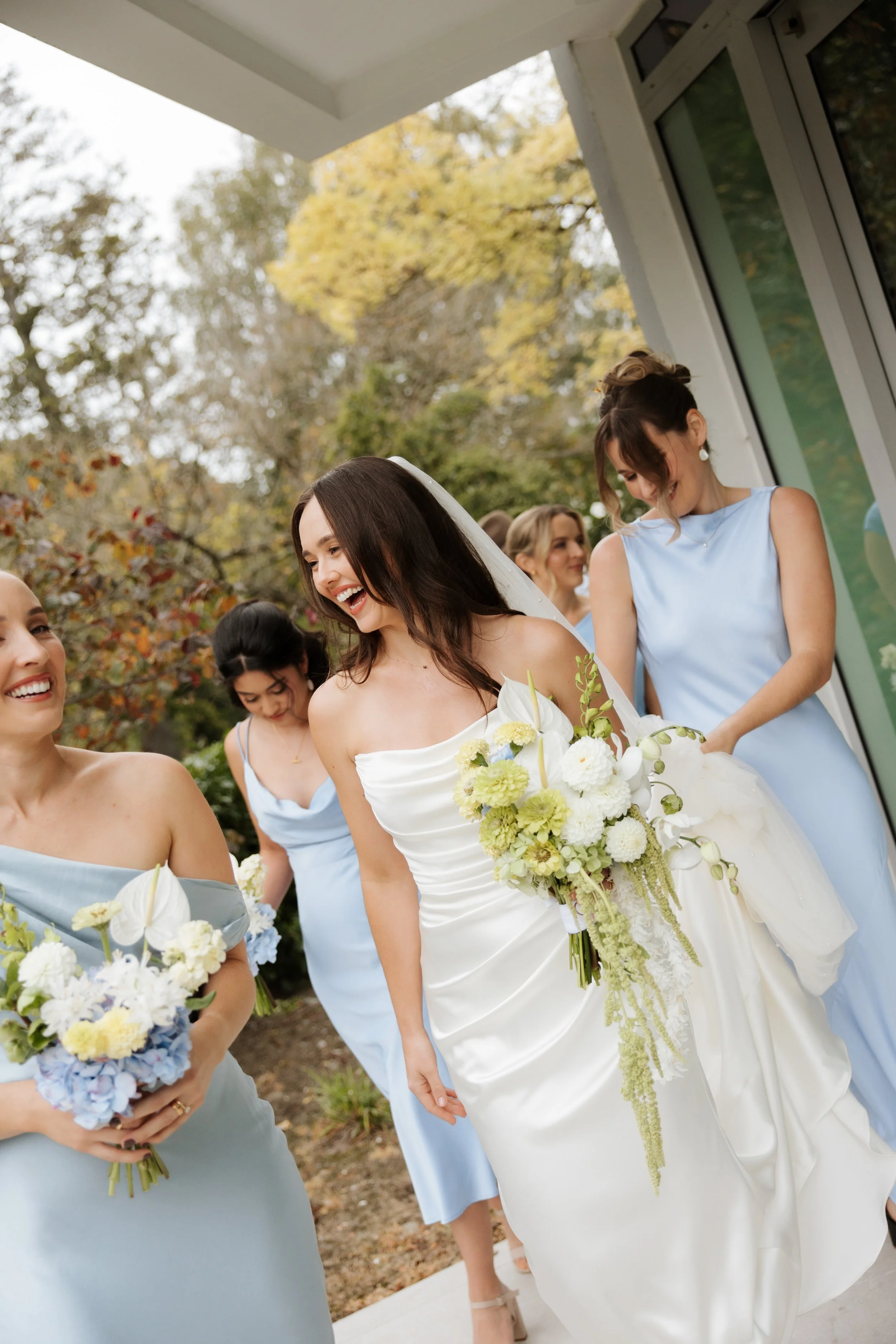 A group of women dressed in light blue and white attire, smiling and holding bouquets of flowers, standing outdoors with trees in the background during a wedding celebration.