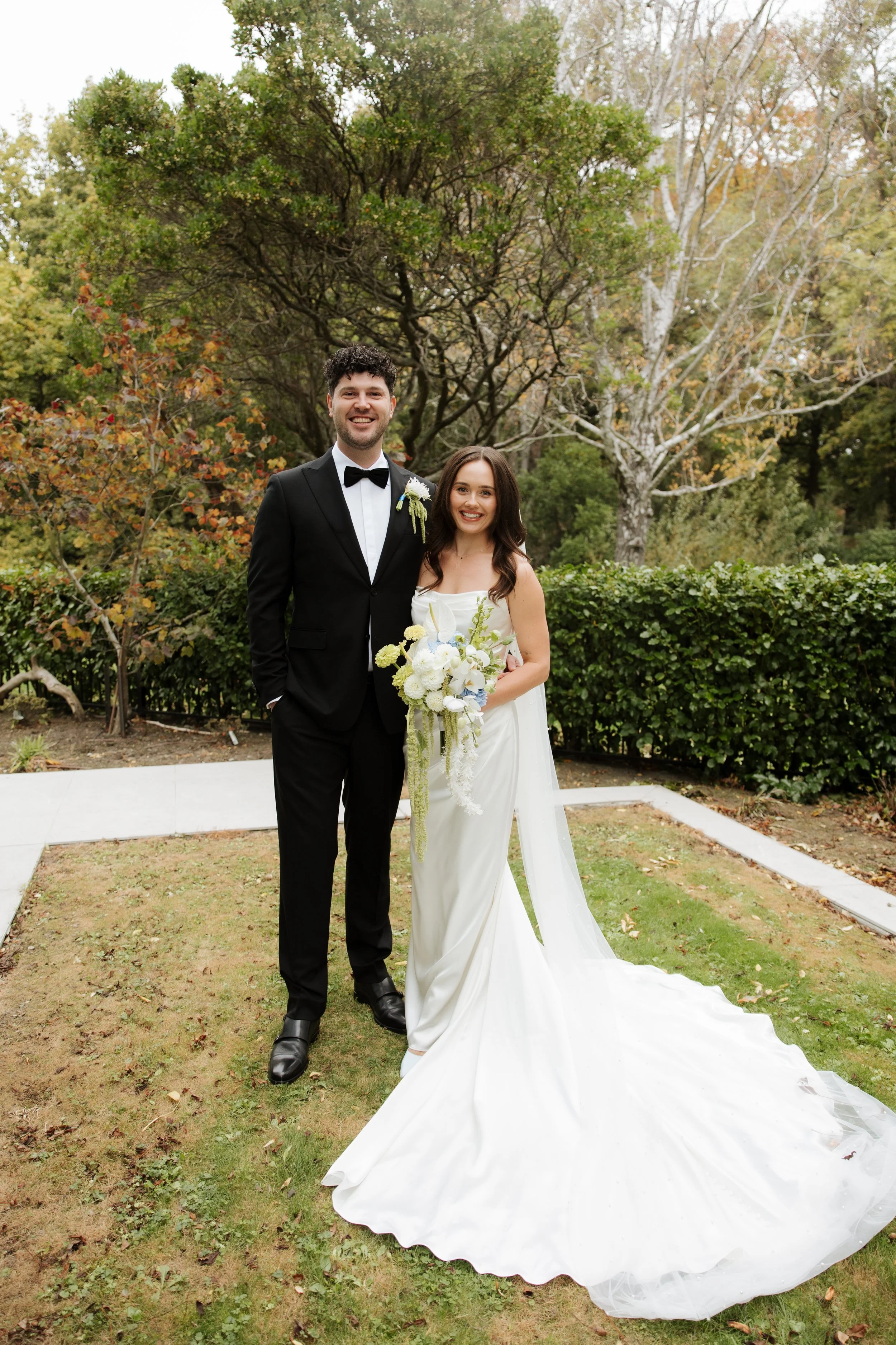 A smiling bride and groom standing outdoors in front of trees, with the bride holding a bouquet of flowers and the groom wearing a black tuxedo with a bowtie.