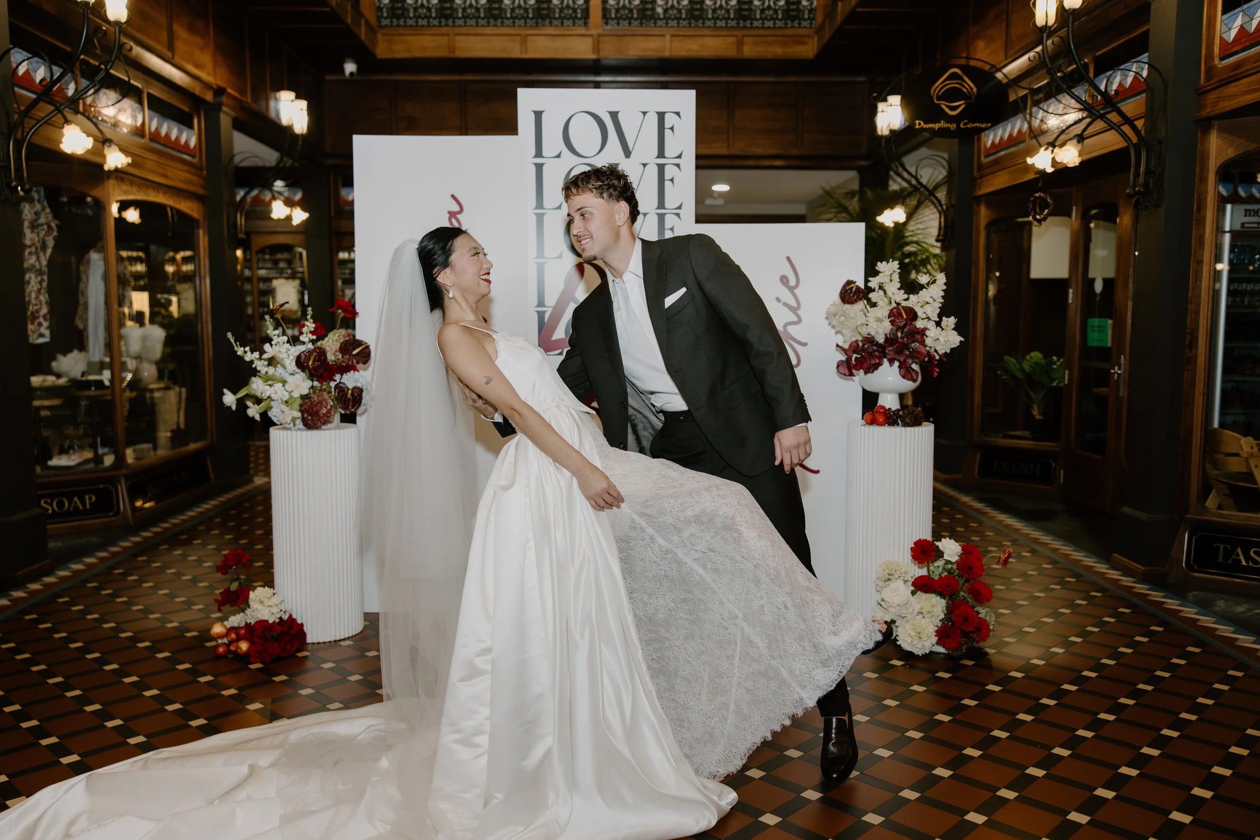 A bride and groom smiling at each other at their wedding reception with love-themed decorations and floral arrangements.