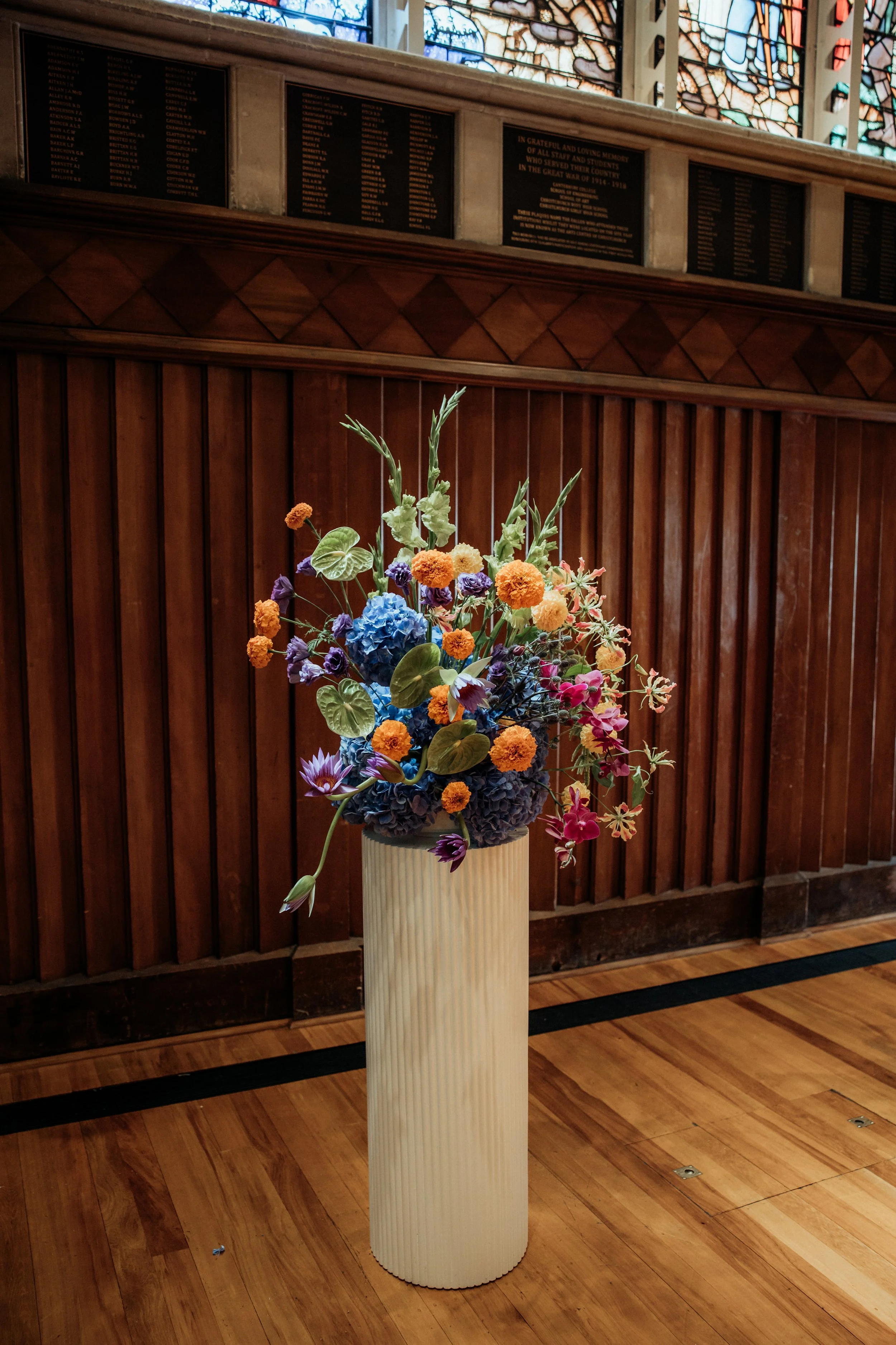 A colorful flower arrangement on a tall, ribbed beige pedestal in front of a wooden-paneled wall with memorial plaques and stained glass windows above.