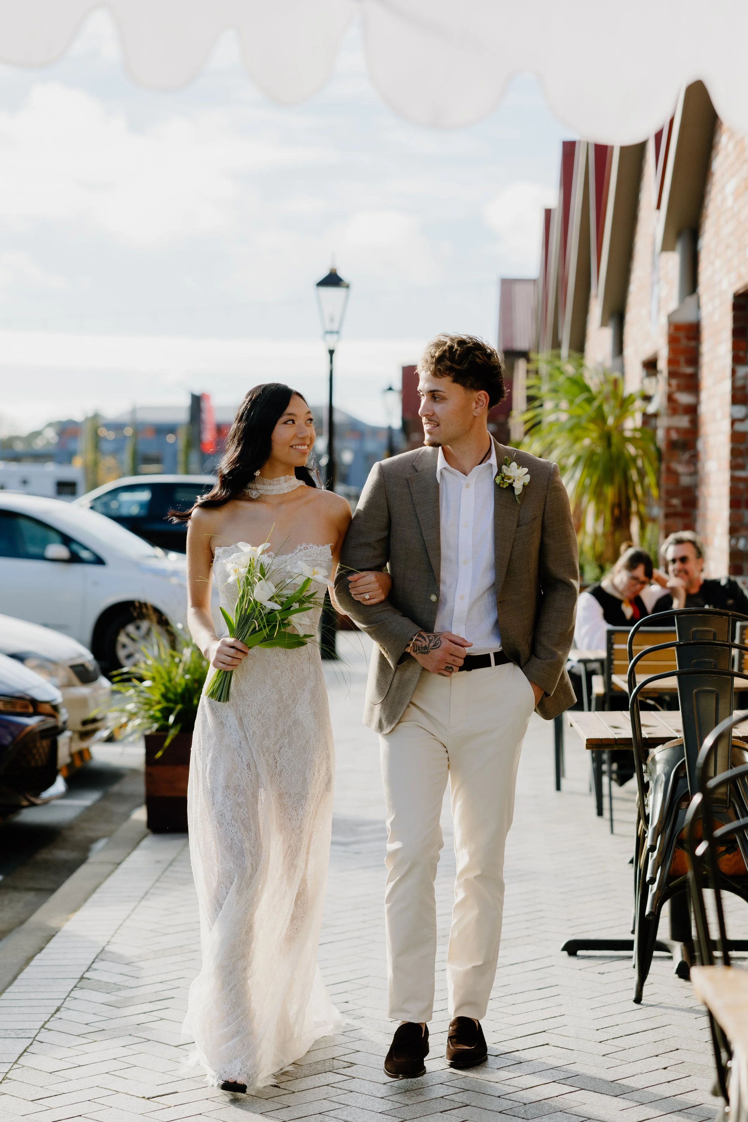 A bride in a white wedding dress holding a bouquet of flowers, and a groom in a gray blazer and white trousers, walking arm-in-arm outdoors during daytime, smiling at each other with a background of cars, a brick building, tables, and people dining.