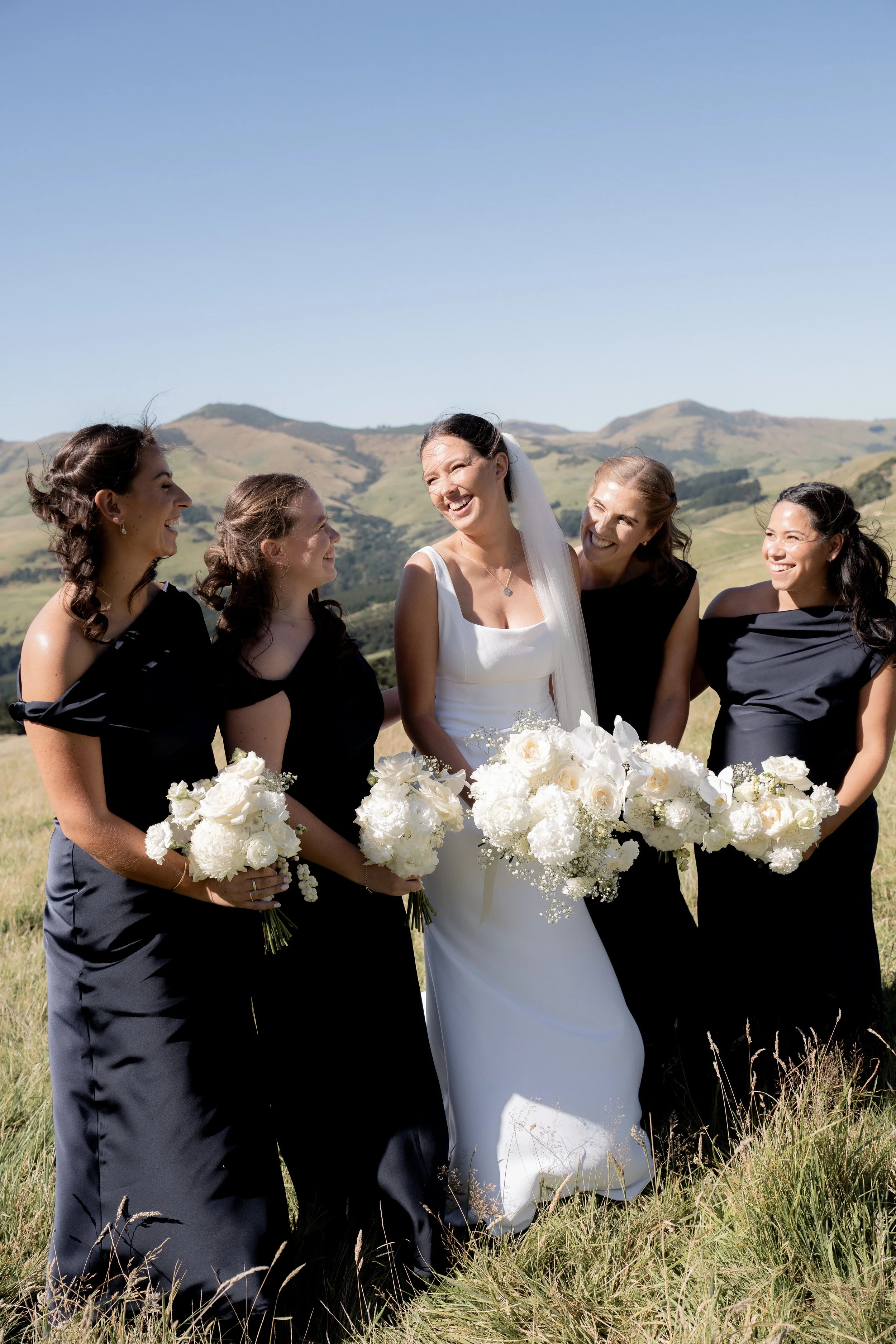 A bride in a white wedding gown standing outdoors with four bridesmaids in black dresses, all holding white bouquets, smiling and laughing together against a backdrop of rolling hills and clear blue sky.