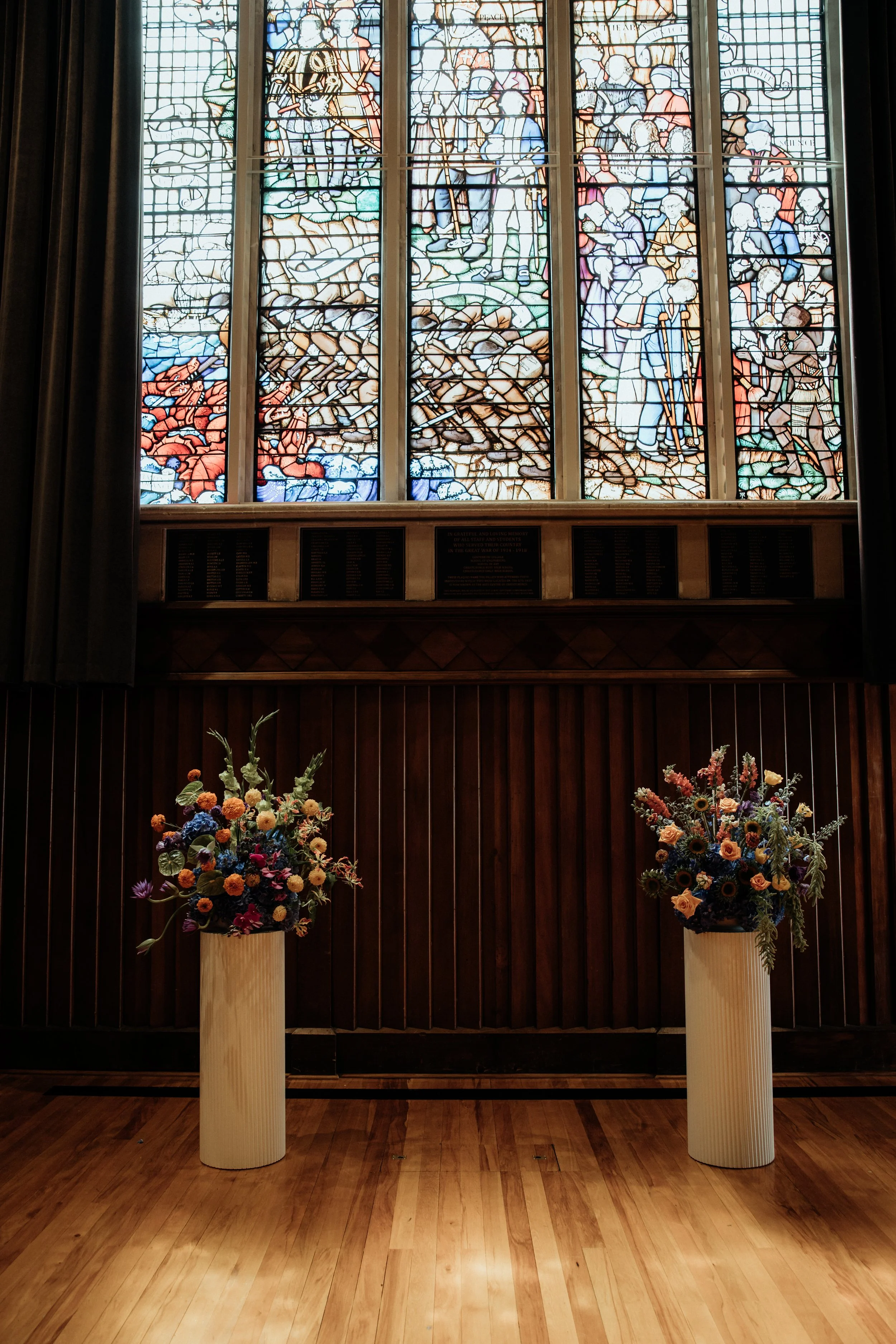 A stained glass window depicting a religious scene, with two large floral arrangements in wooden vases placed in front of a dark wooden wall.