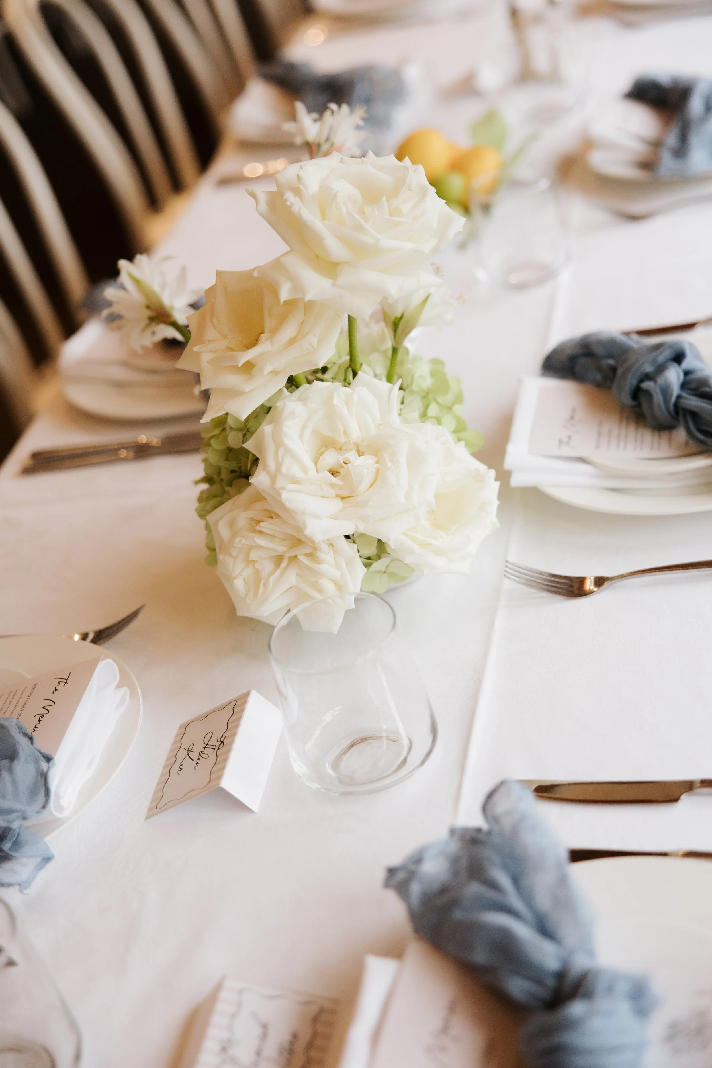 A table setting with a centerpiece of white roses and other flowers, place cards, napkins tied with blue fabric, plates, and silverware.