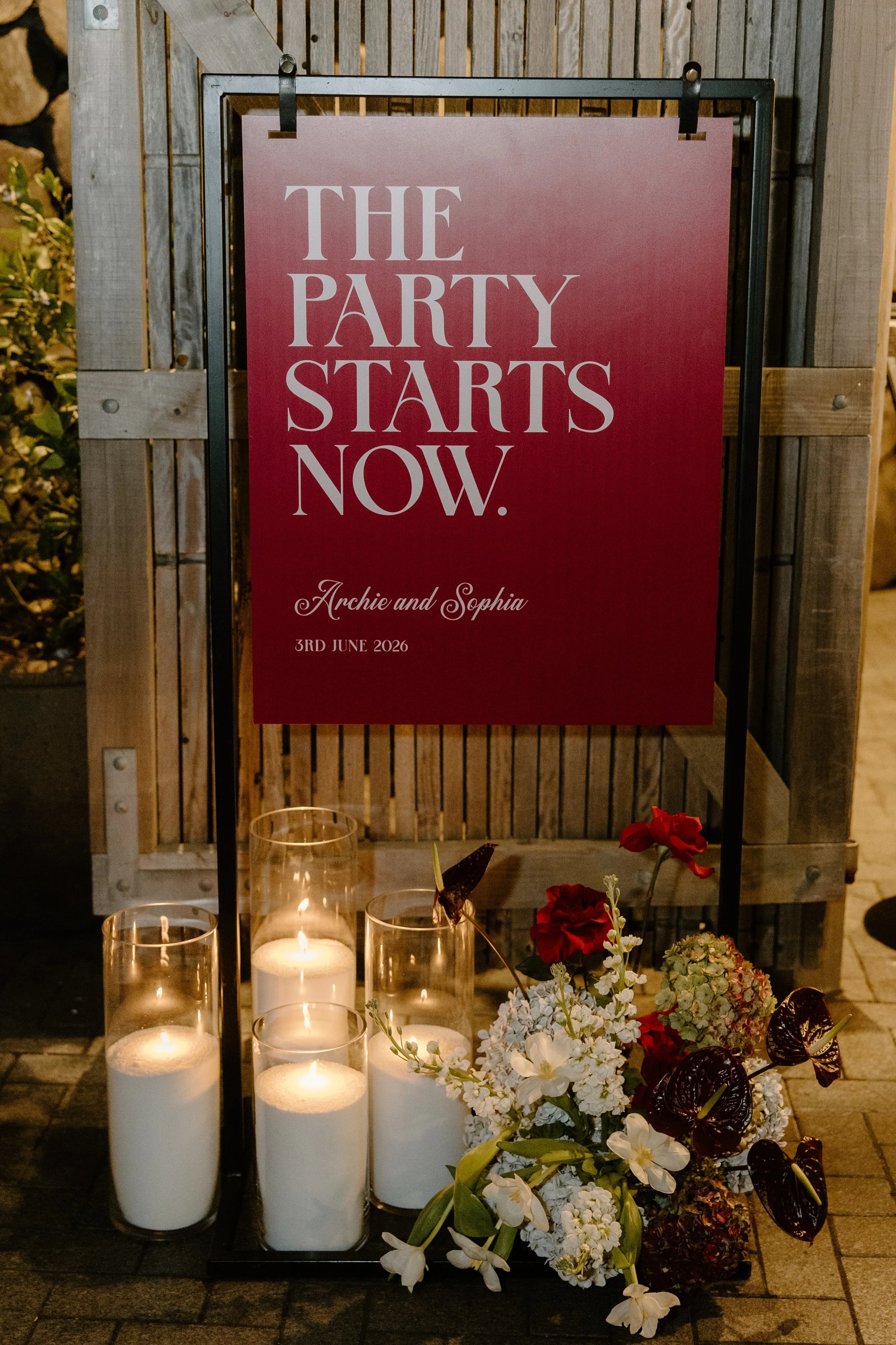 A sign that reads 'The Party Starts Now' with following names, Archie and Sophia, and the date '3rd June 2026' in elegant script. The sign is surrounded by illuminated candles in tall glass holders and floral arrangements, placed outdoors against a w