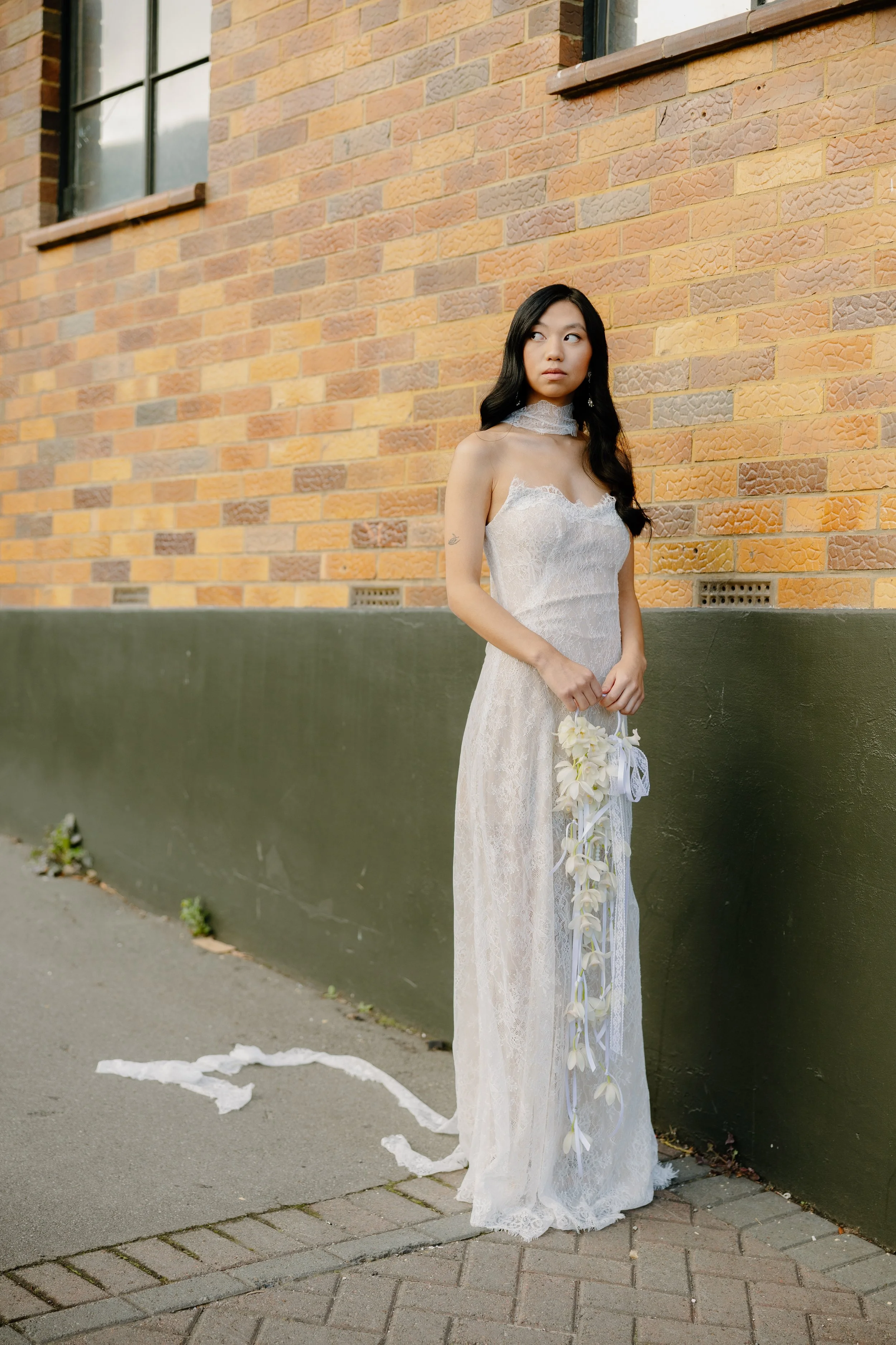 A woman in a white lace wedding dress holding a bouquet of flowers stands outside against a brick wall with windows.