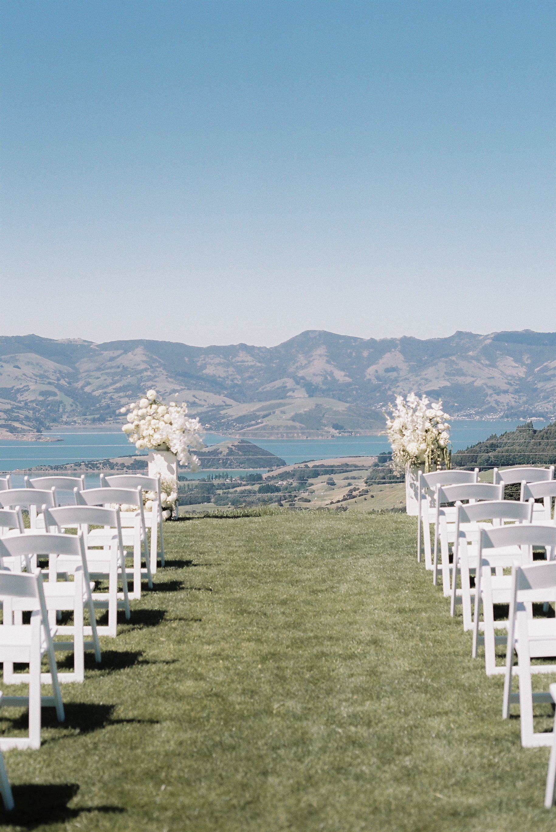 Outdoor wedding setup with white chairs on green grass, decorated with white flower arrangements, overlooking a scenic lake and mountain backdrop.