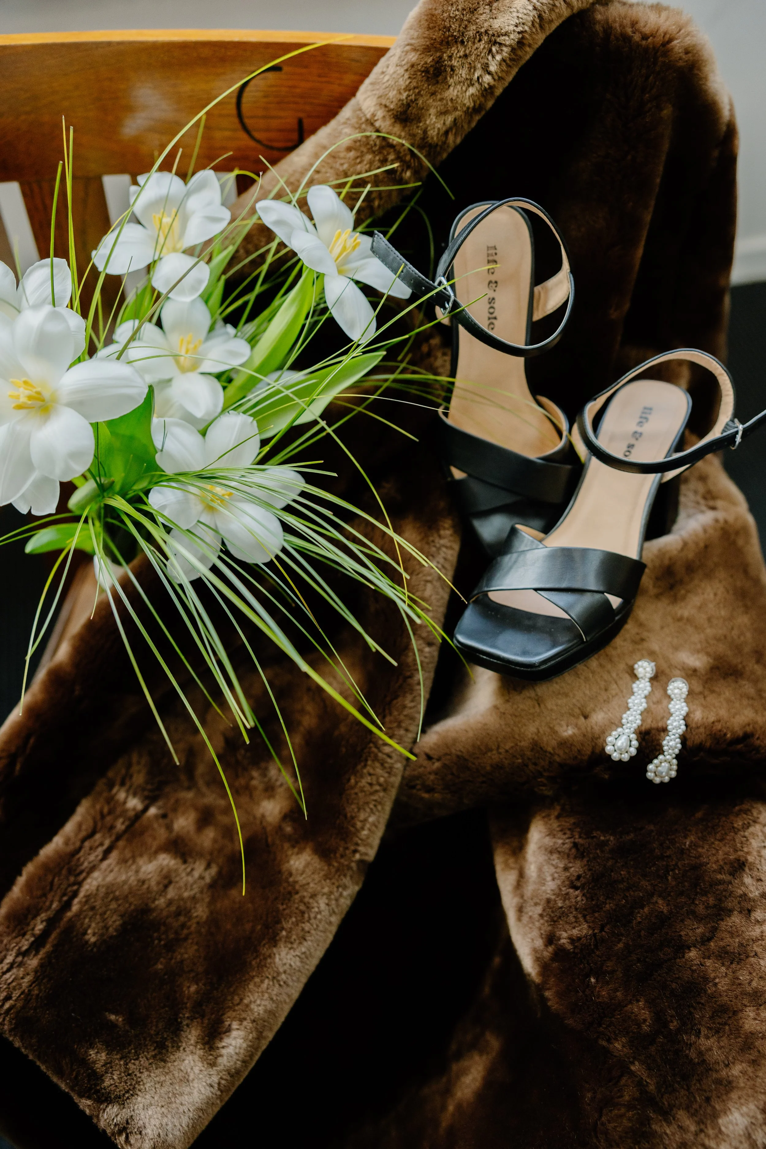 Black high-heeled sandals with ankle straps placed on a brown faux fur fabric, next to a bouquet of white flowers with yellow centers, and a pair of pearl earrings.