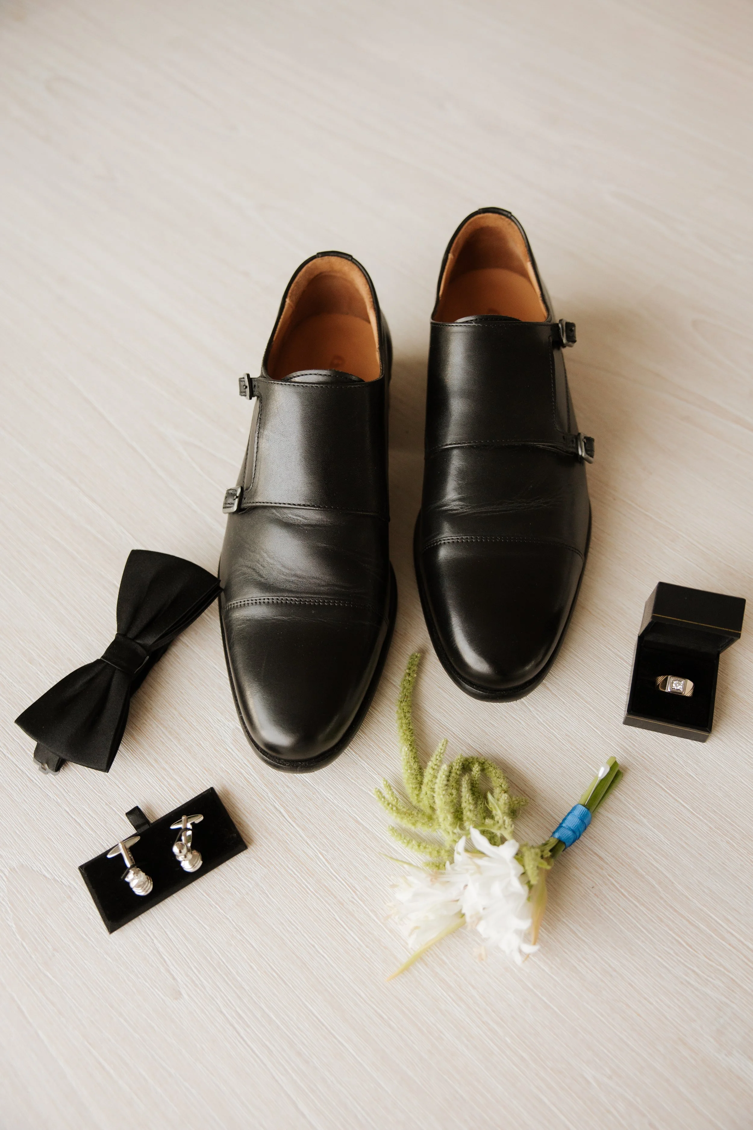 Pair of black formal men's shoes, black bow tie, black cufflinks, a decorative ring, some flowers and greenery, and other jewelry on a light wooden surface.