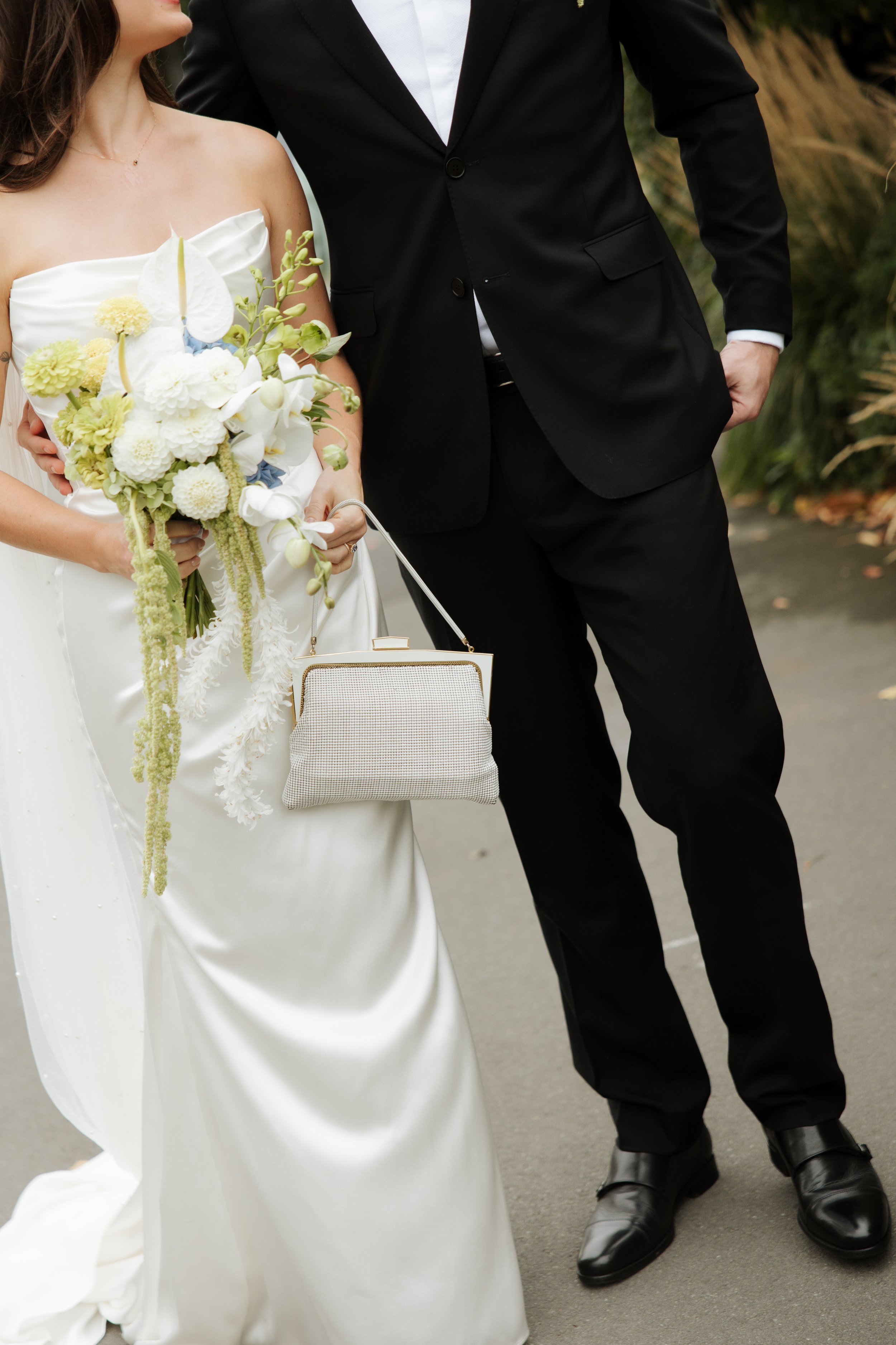 A bride in a white off-shoulder wedding gown holding a bouquet of white, pale yellow, and pastel green flowers, standing next to a groom in a black suit and white shirt, with a handbag hanging from her wrist. The groom has one hand in his pocket, and