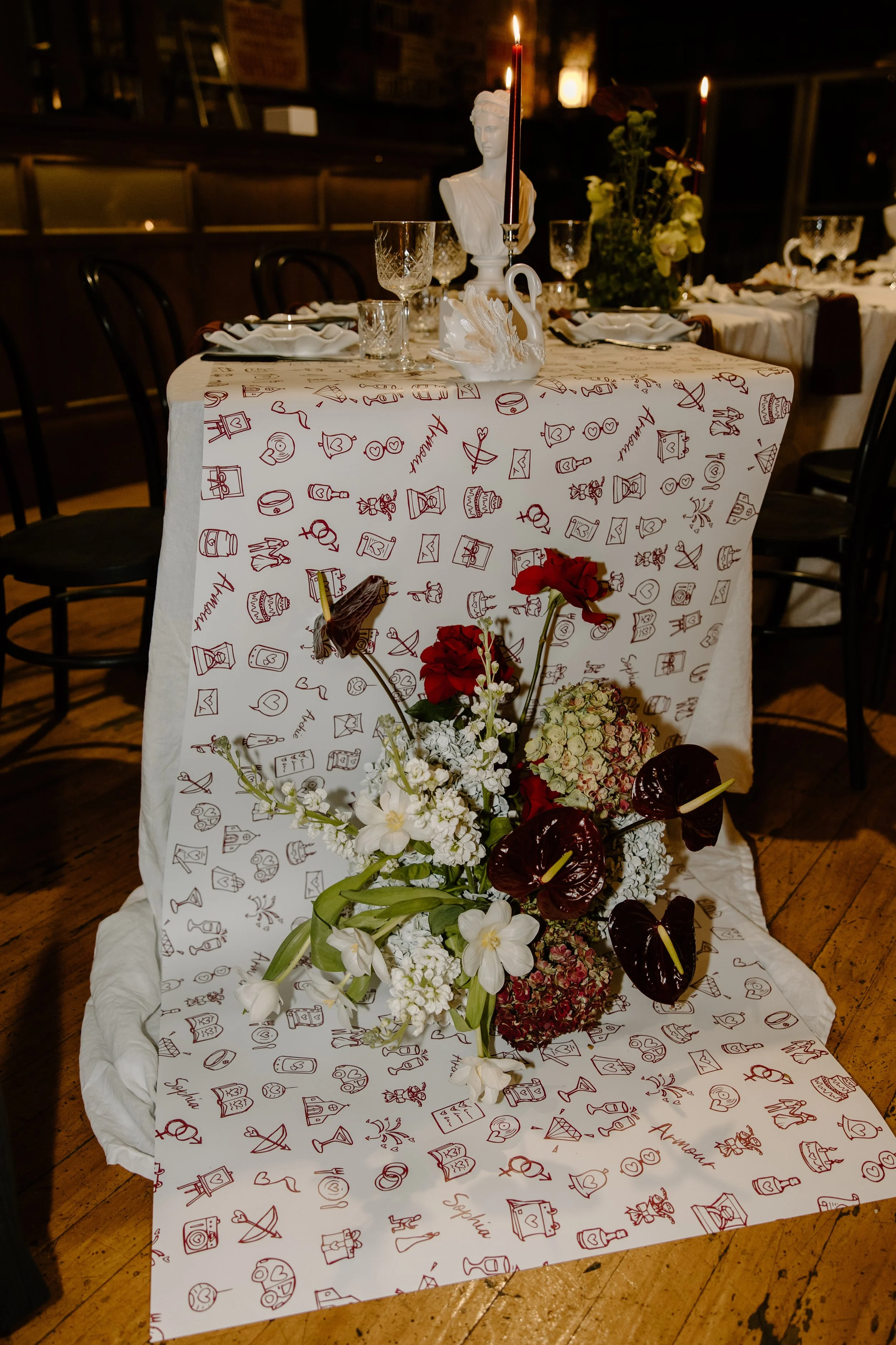 A decorated table with a white tablecloth featuring red doodles and signatures, floral arrangements, candlesticks, and decorative items in a dimly lit setting.