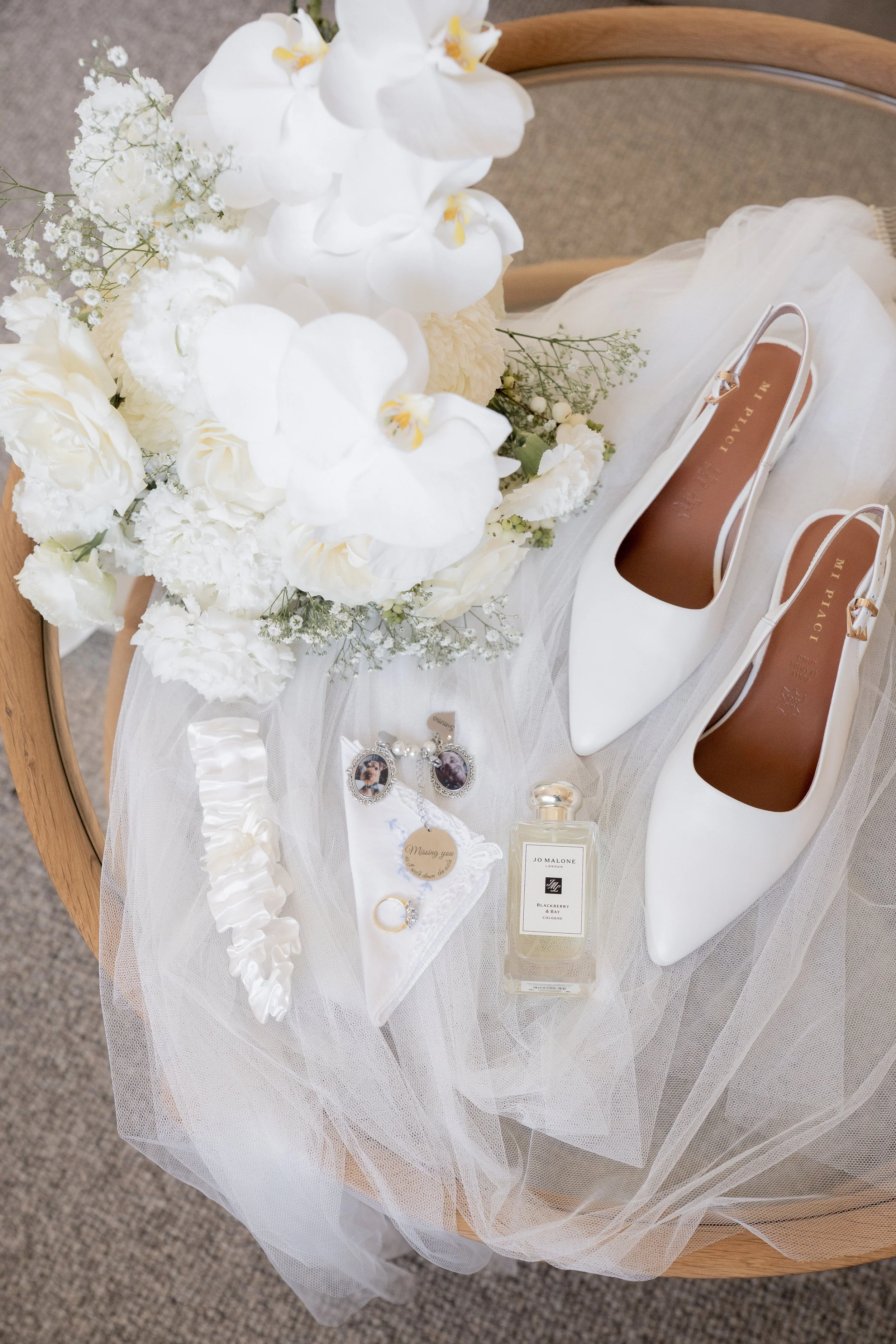 A wedding bouquet of white flowers, featuring orchids, roses, and baby's breath, resting on tulle fabric. Beside it are white high-heeled shoes marked 'In Pai' and a selection of jewelry, perfume, and accessories on a round wooden chair.