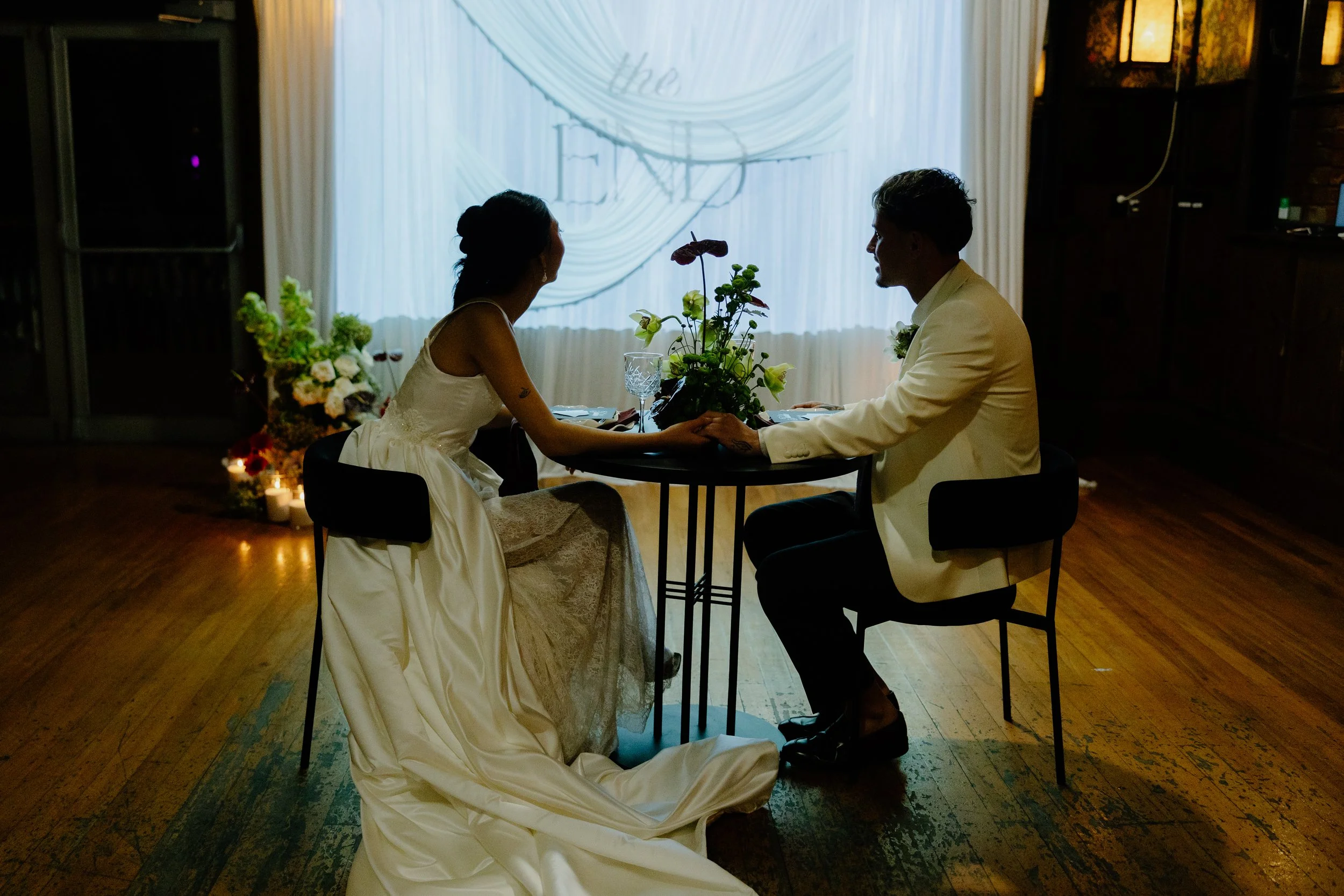 A bride and groom sitting at a table holding hands, in a dimly lit indoor setting decorated with flowers and candles, with a curtain in the background.