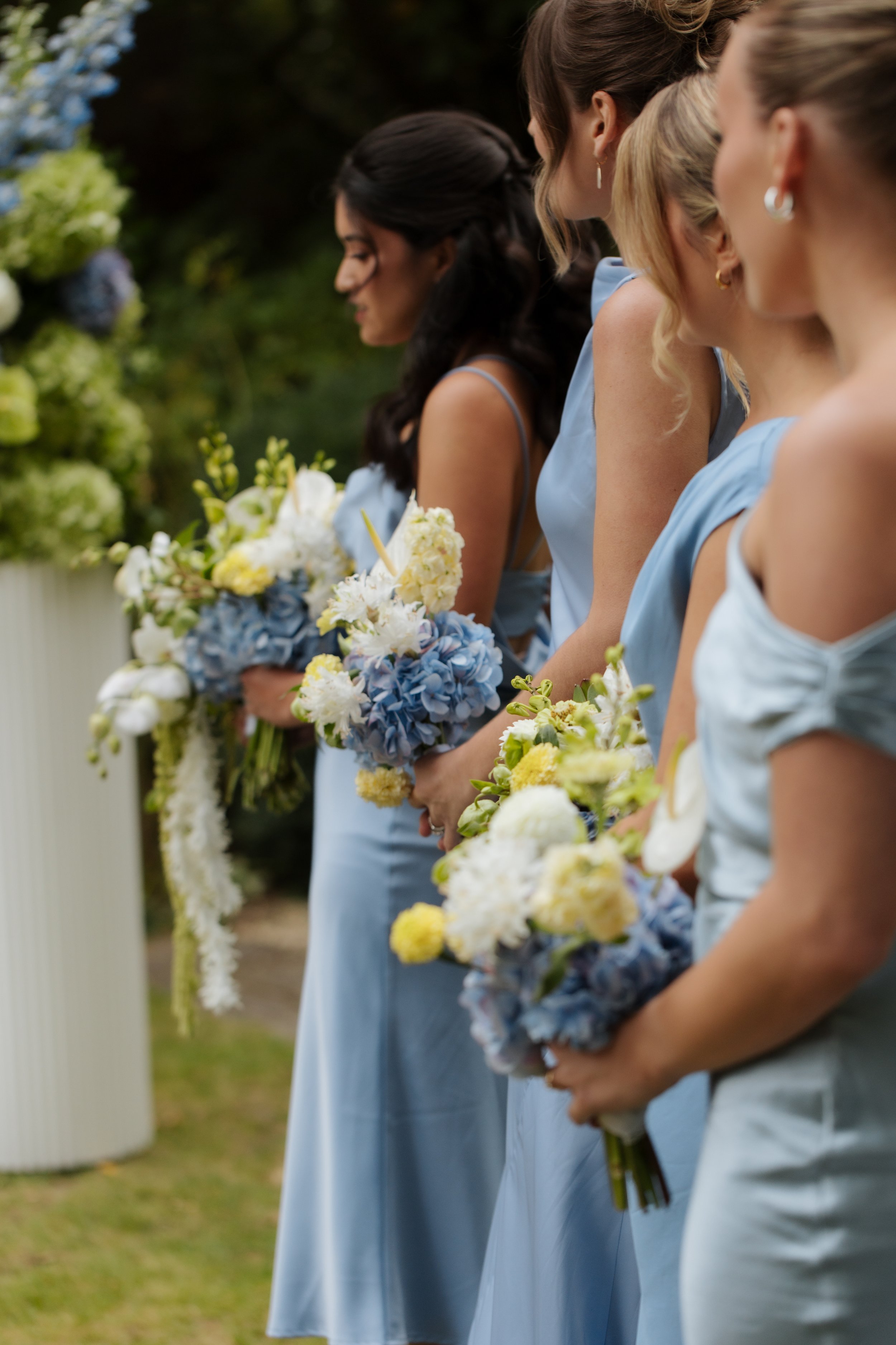 Line of women wearing light blue dresses holding bouquets, likely at a wedding or formal outdoor event.