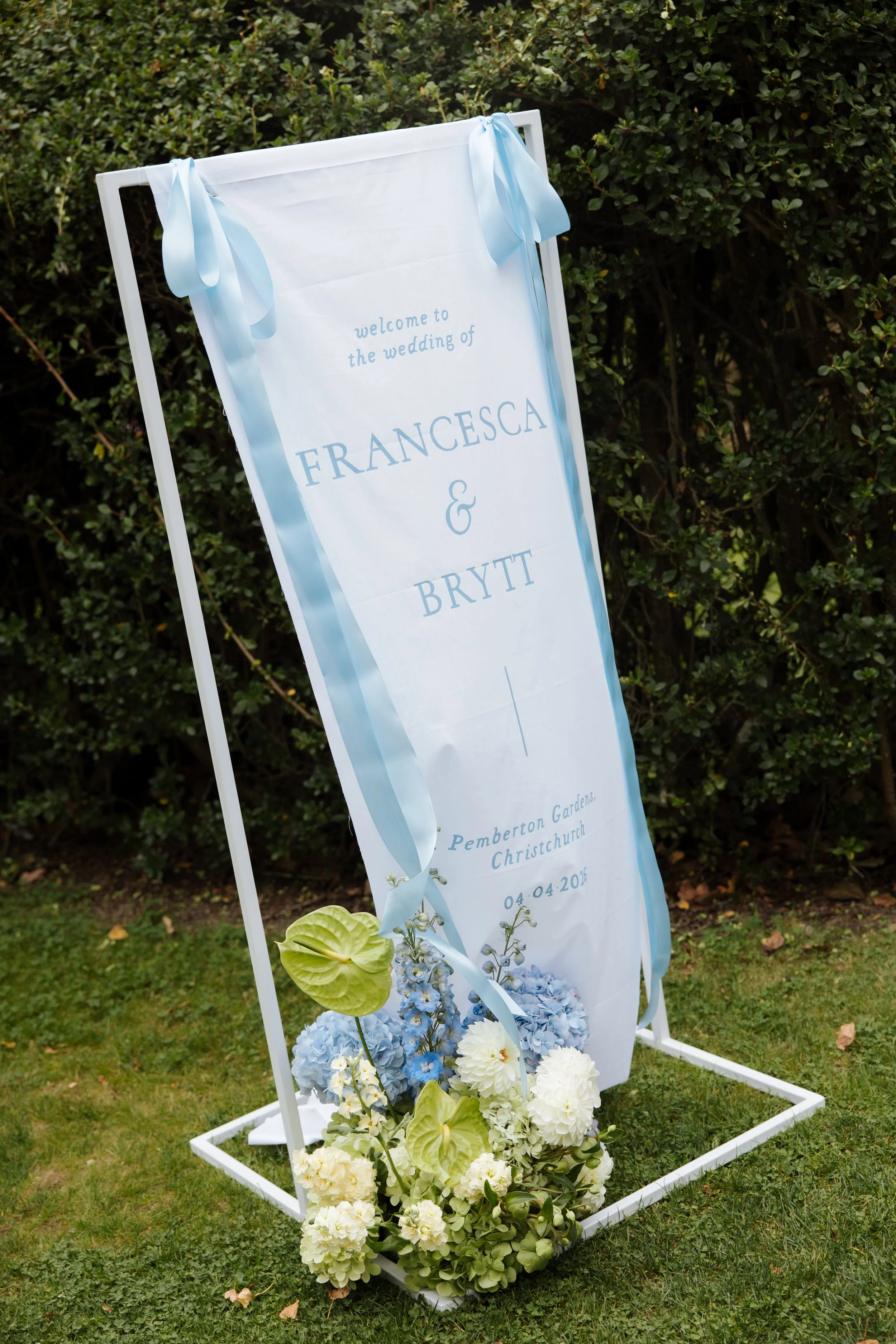 Wedding welcome sign with blue and white flowers, light blue ribbons, and a white frame, placed outdoors on grass, with green bushes in the background.