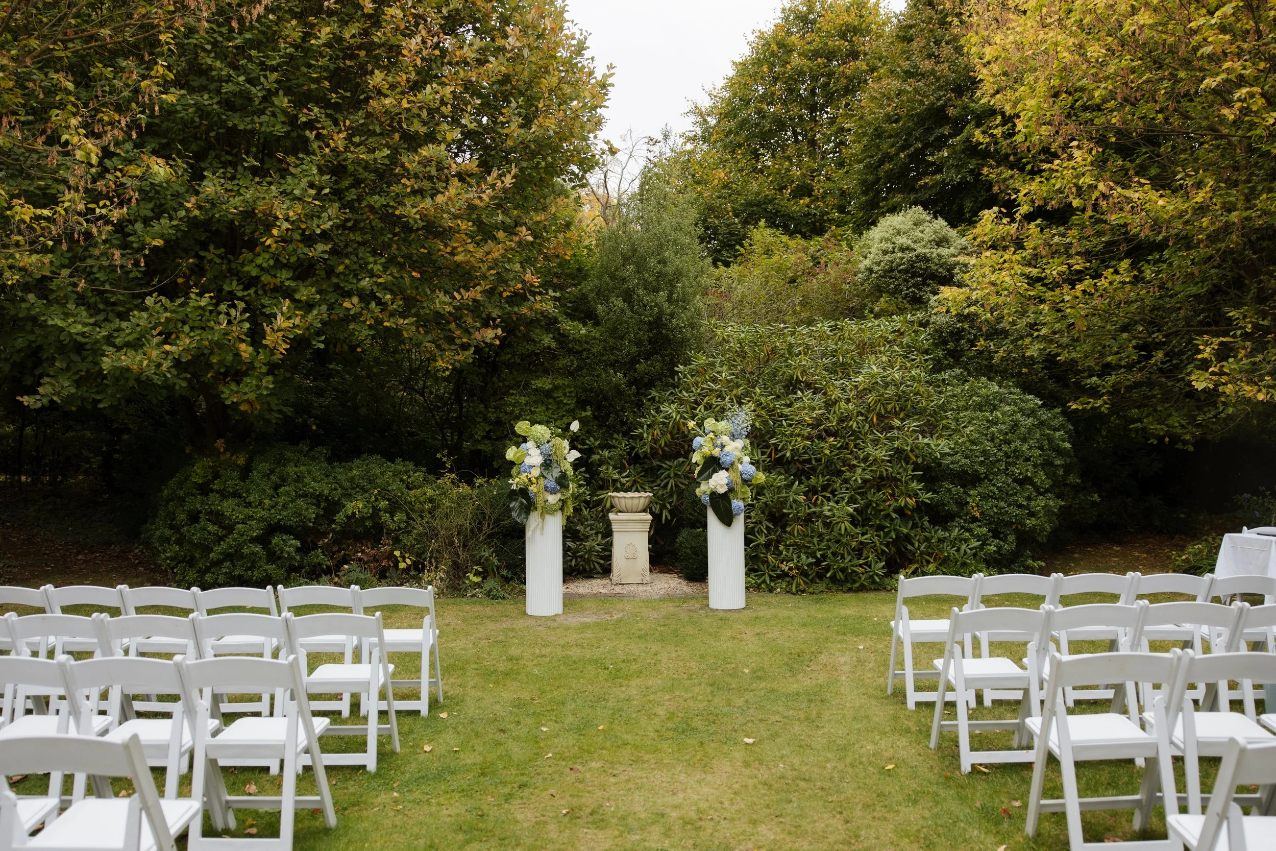 Outdoor wedding ceremony setup with rows of white chairs facing an altar with white columns and floral arrangements, surrounded by lush green trees.