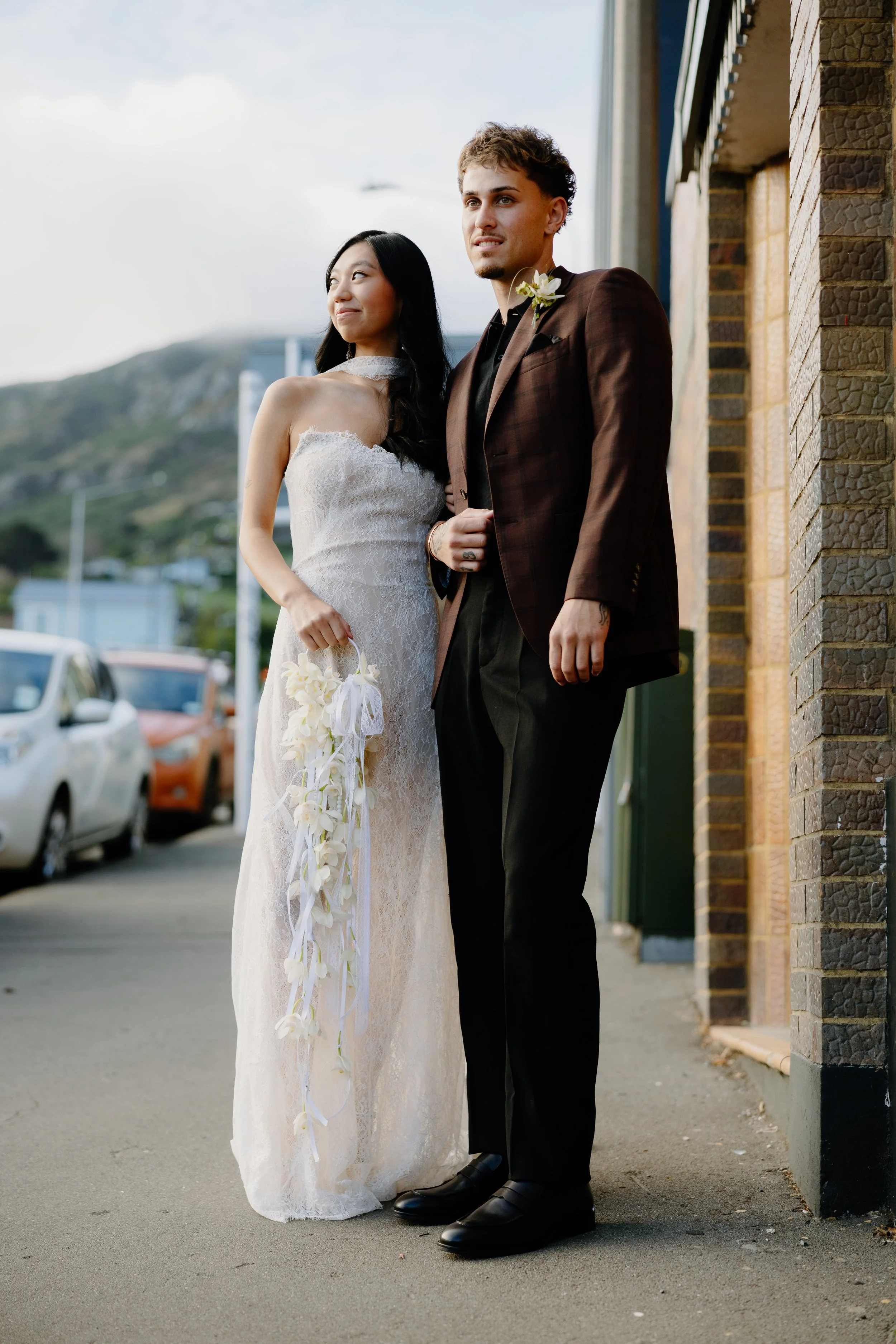 A bride and groom standing side by side on a city sidewalk, dressed in wedding attire. The bride wears a white lace wedding gown and holds a bouquet of white flowers, smiling softly. The groom wears a dark plaid suit with a white boutonnière and look