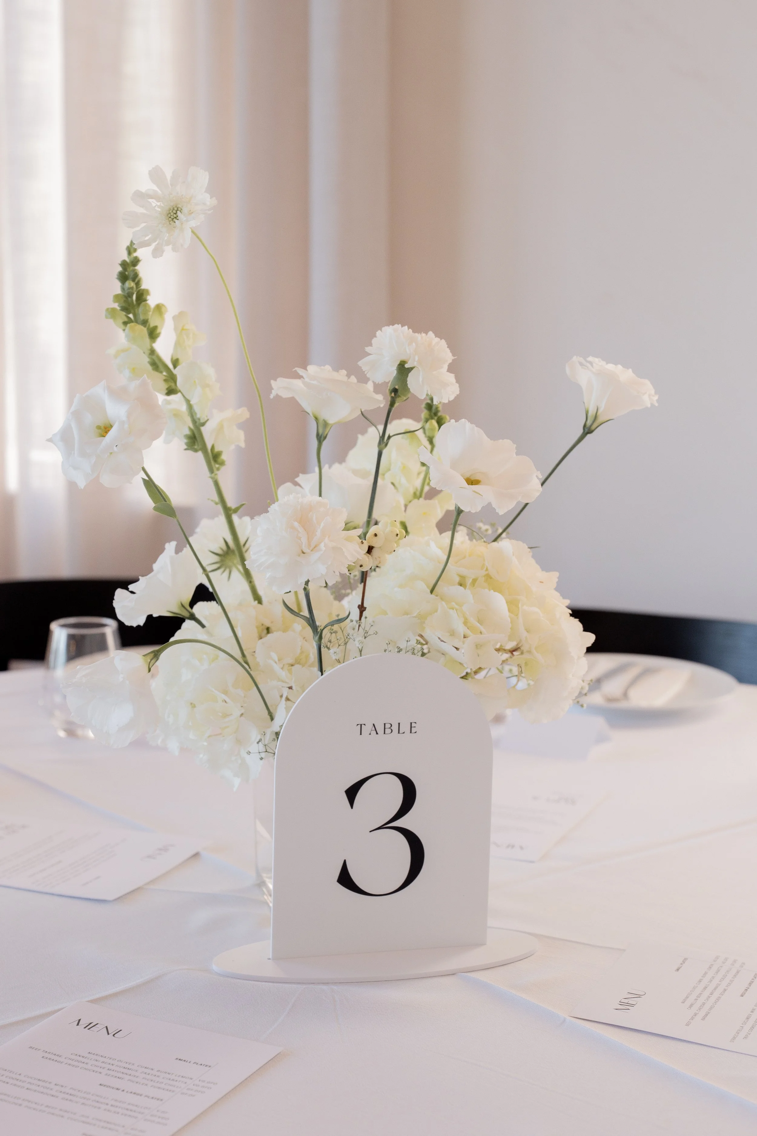 Elegant table centerpiece with white flowers, including lisianthus and delphiniums, on a white table with a table number '3' card. In the background, there are menu cards and a clear glass, suggesting a formal event or wedding reception.