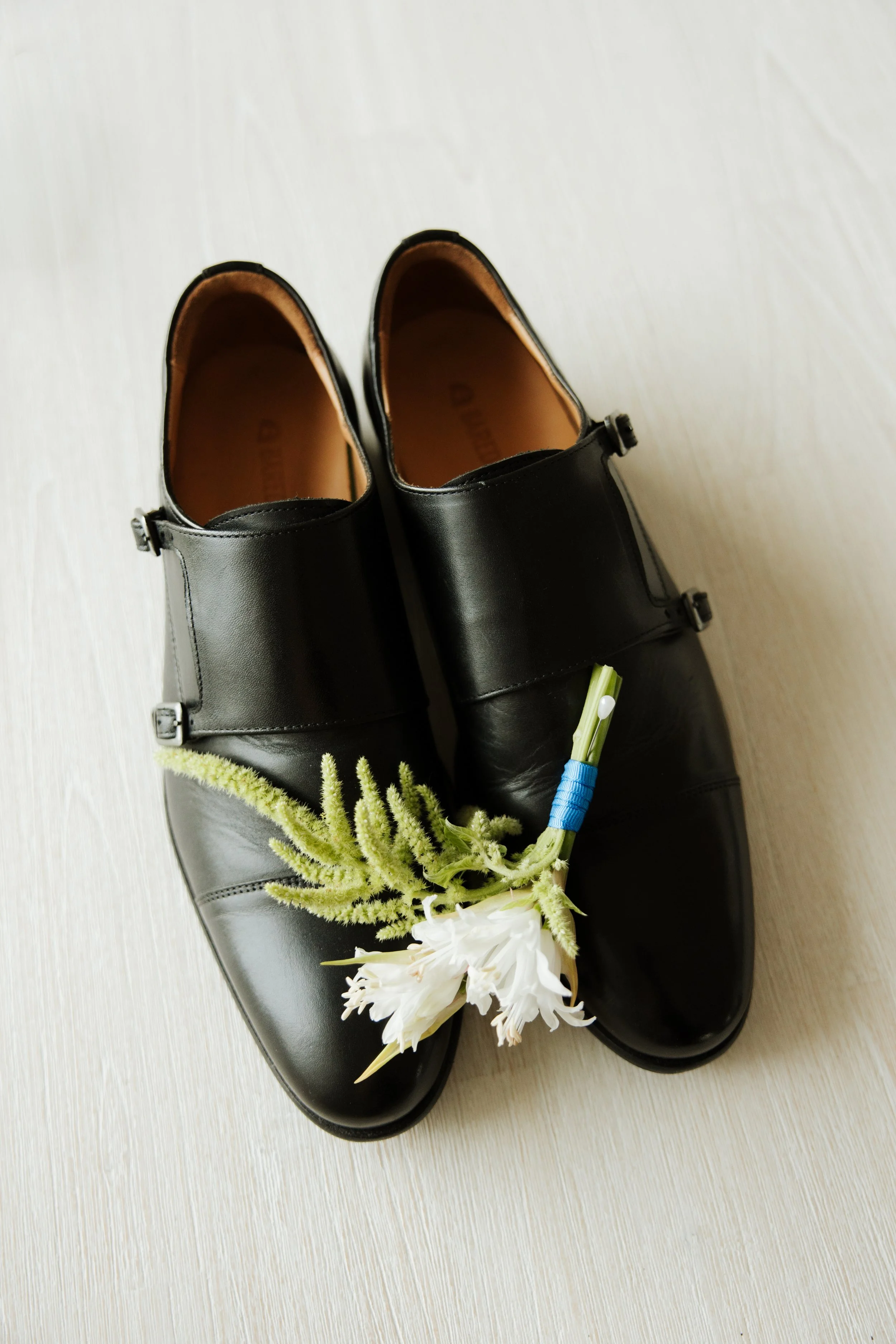 Pair of black leather shoes with white flowers and green foliage laid on top, placed on a light-colored wood floor.