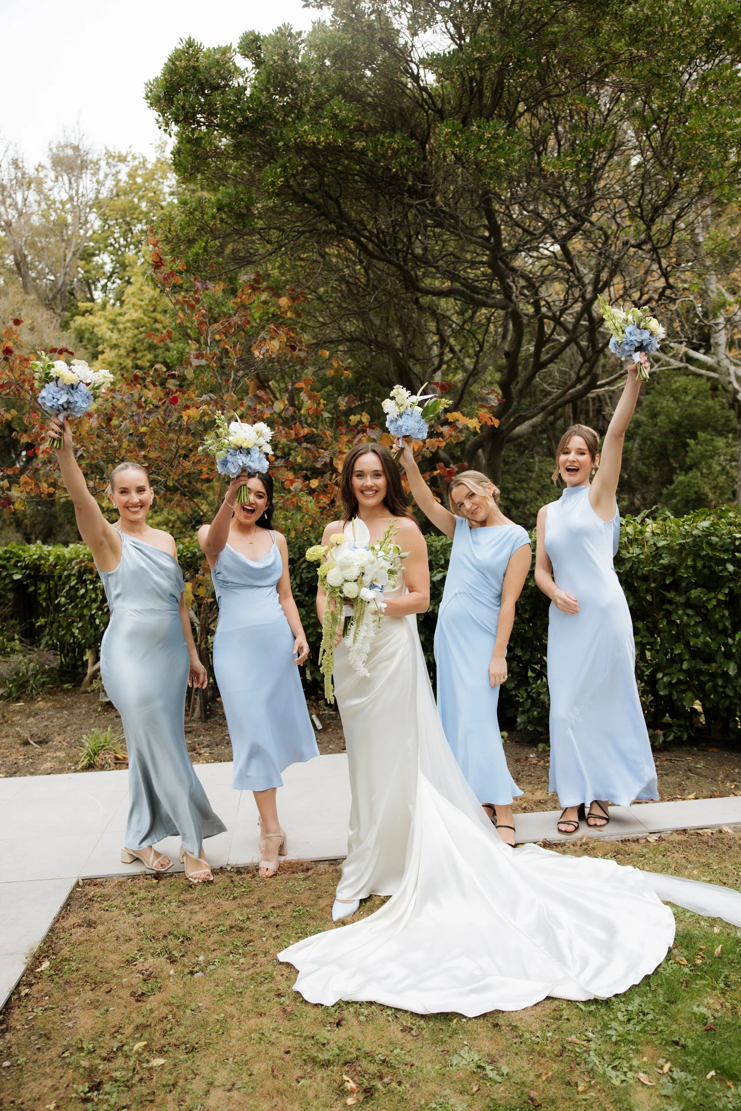 A bride in a white wedding dress surrounded by five bridesmaids in light blue dresses outside in a garden. The bridesmaids are holding bouquets and raising their arms in celebration. The bride is holding a bouquet of white flowers, smiling.