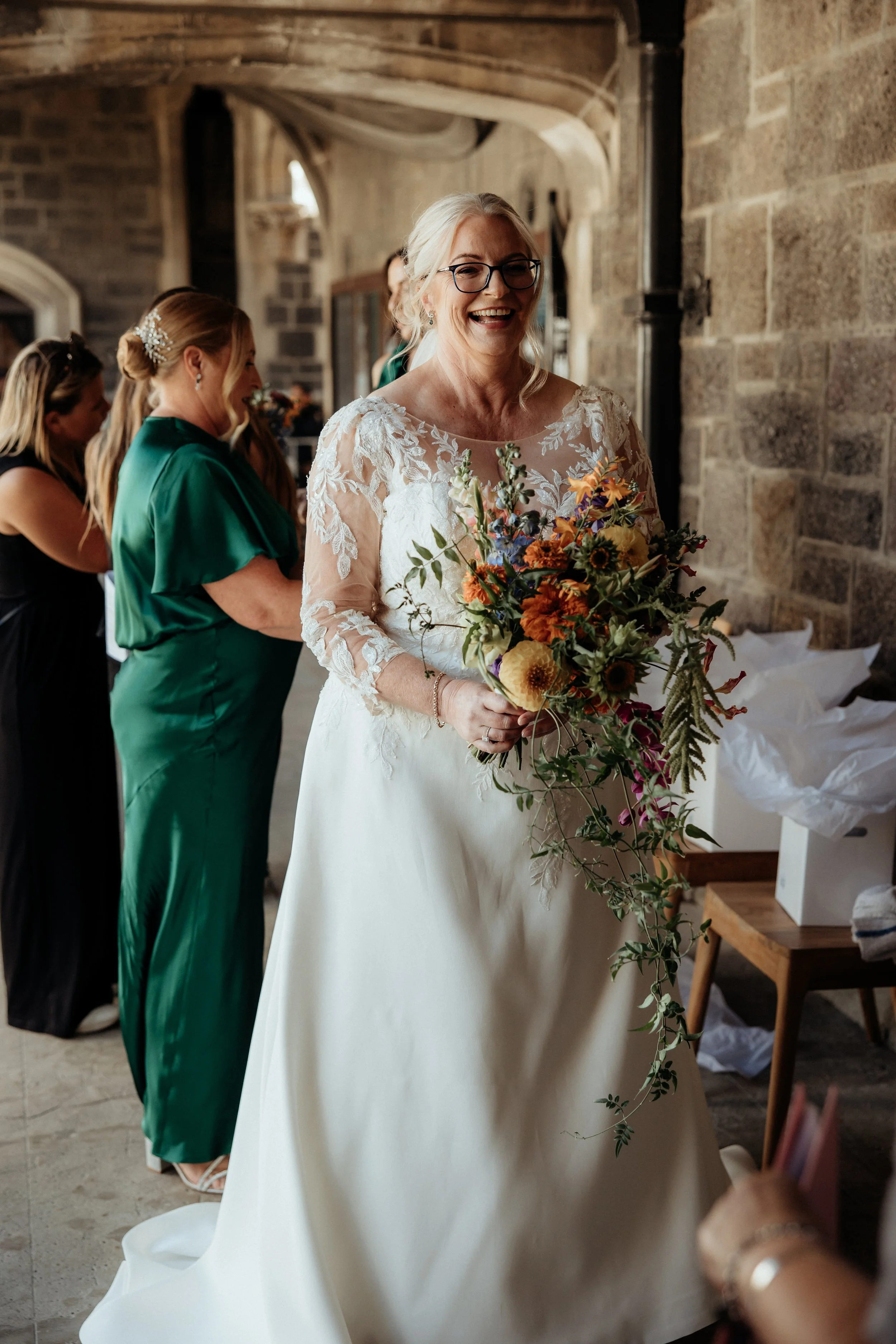 A smiling bride holding a bouquet of colorful flowers, wearing a lace wedding dress with long sleeves, standing in a stone-walled church. Several women, including a bridesmaid in a green dress, are in the background.