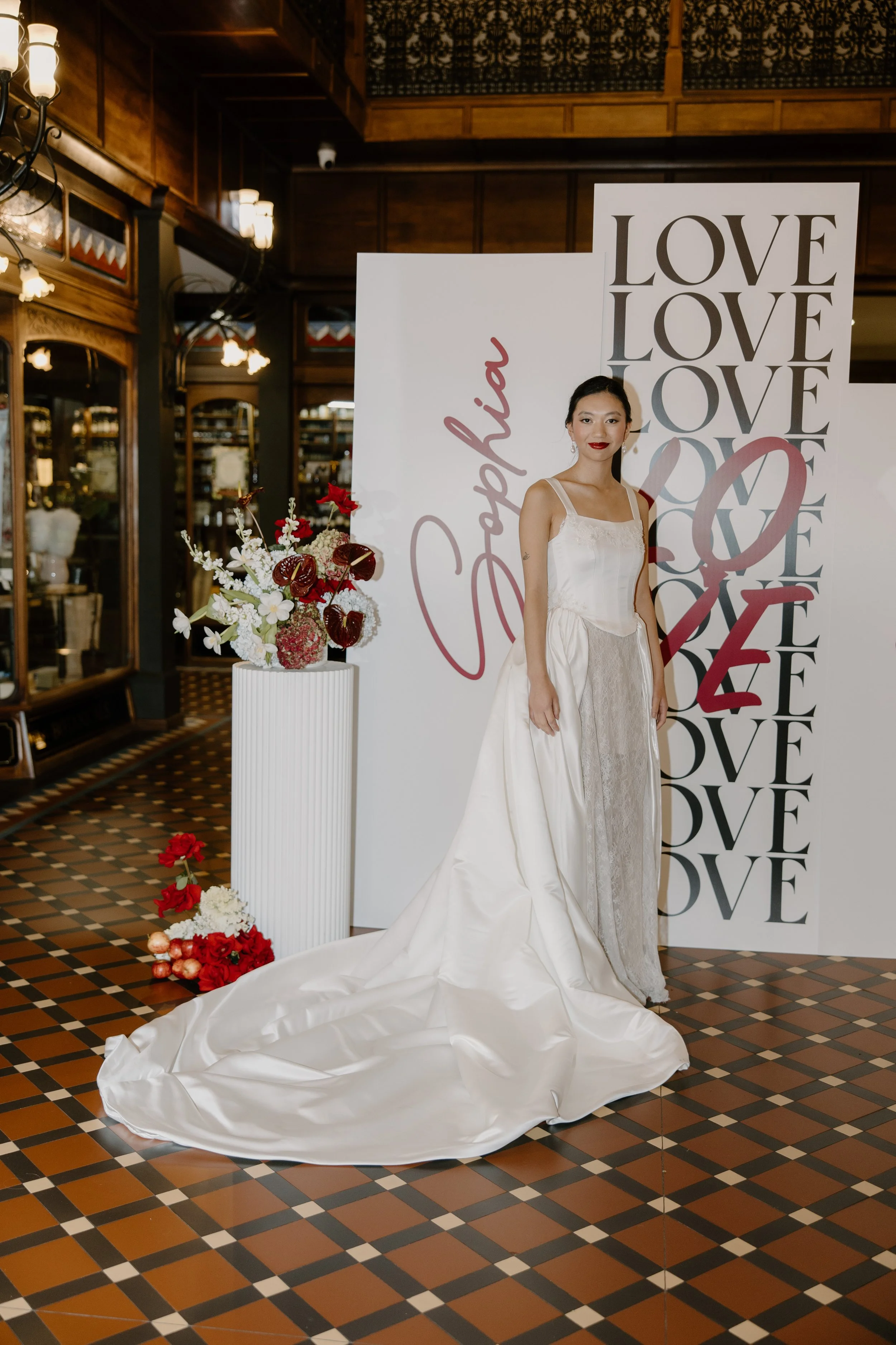 A woman in a white wedding dress standing in front of a backdrop with the words 'LOVE' repeated multiple times, at a wedding or bridal event. There is a white floral arrangement with red and white flowers on a white pedestal and some flowers and deco