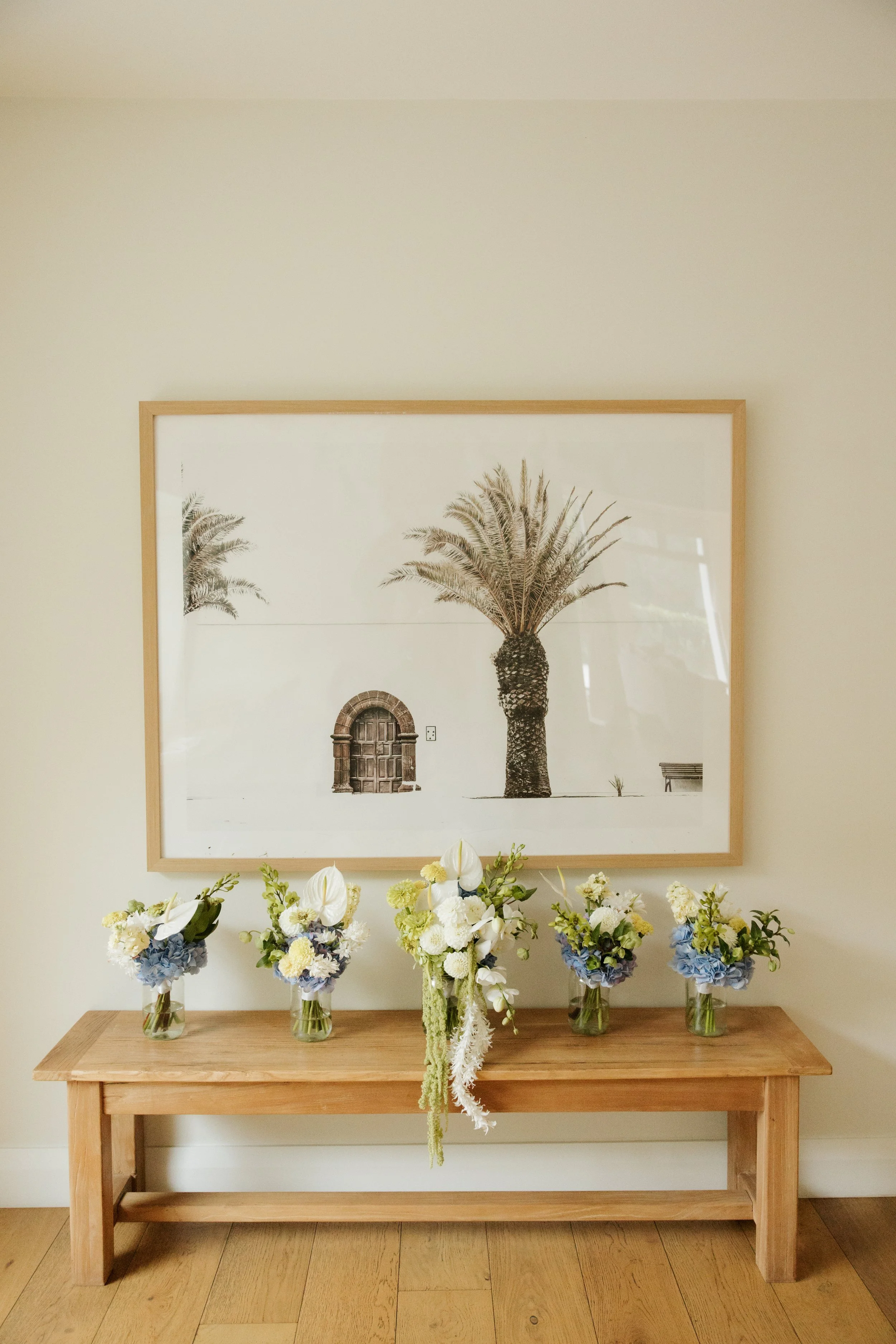 Wooden table with five vases of flowers, a framed black and white picture of a palm tree and an arched door, and a floral arrangement hanging over the table.