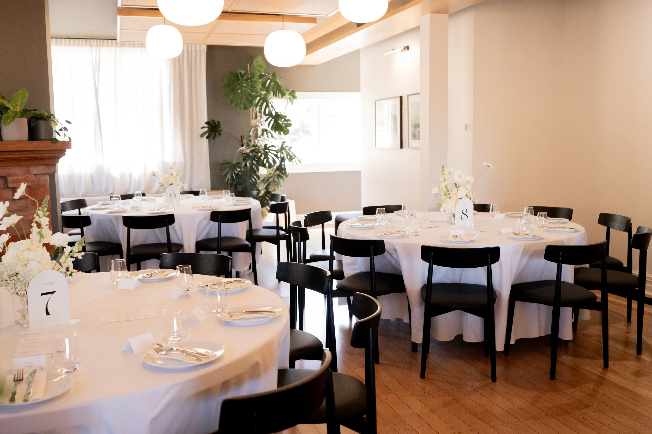 A dining area set up for a formal event with round tables covered in white tablecloths, black chairs, floral centerpieces, and place settings including glasses, silverware, and napkins. Large windows with white curtains illuminate the room. There are