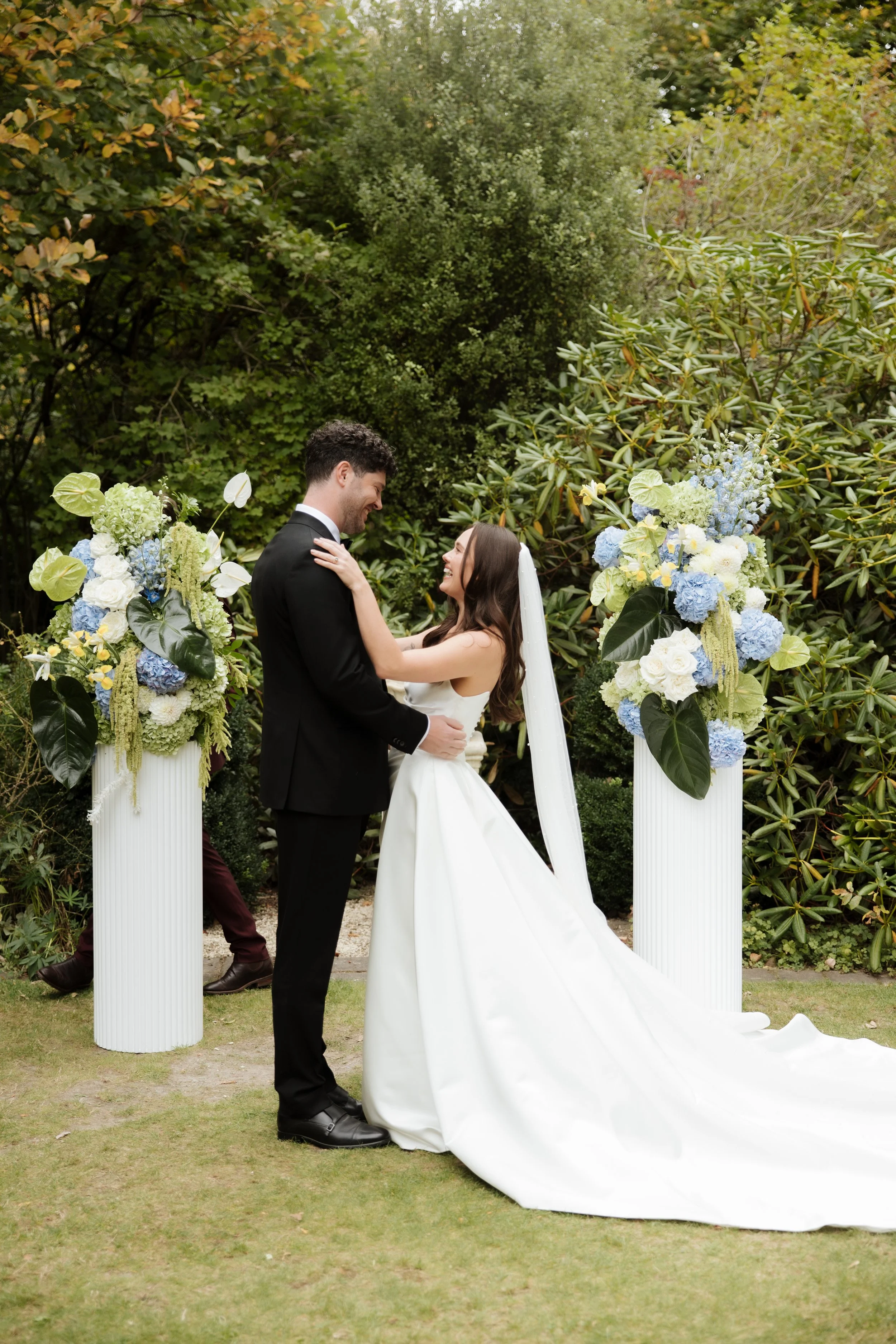 A bride and groom stand together outdoors amidst greenery, smiling and looking at each other during their wedding ceremony, with floral decorations on white columns surrounding them.
