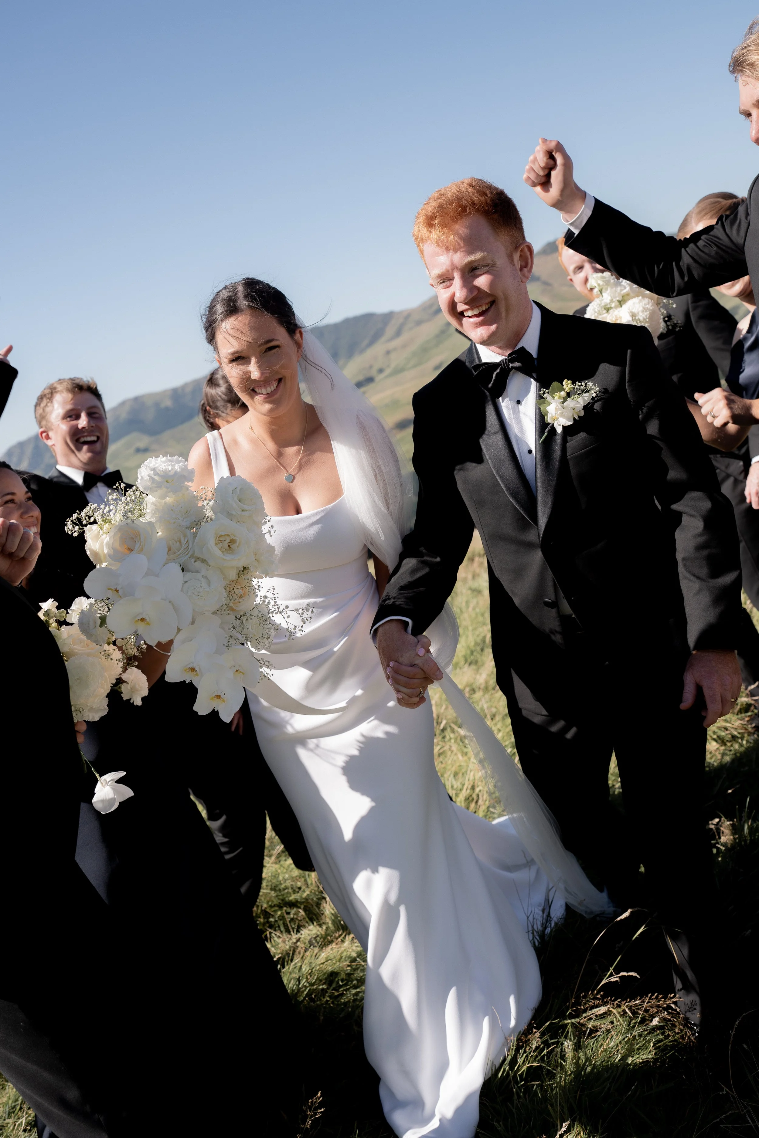 A bride and groom with wedding guests outdoors on a sunny day, mountains in the background. The bride is wearing a white wedding dress and holding a bouquet of white flowers. The groom is in a black tuxedo and smiling.