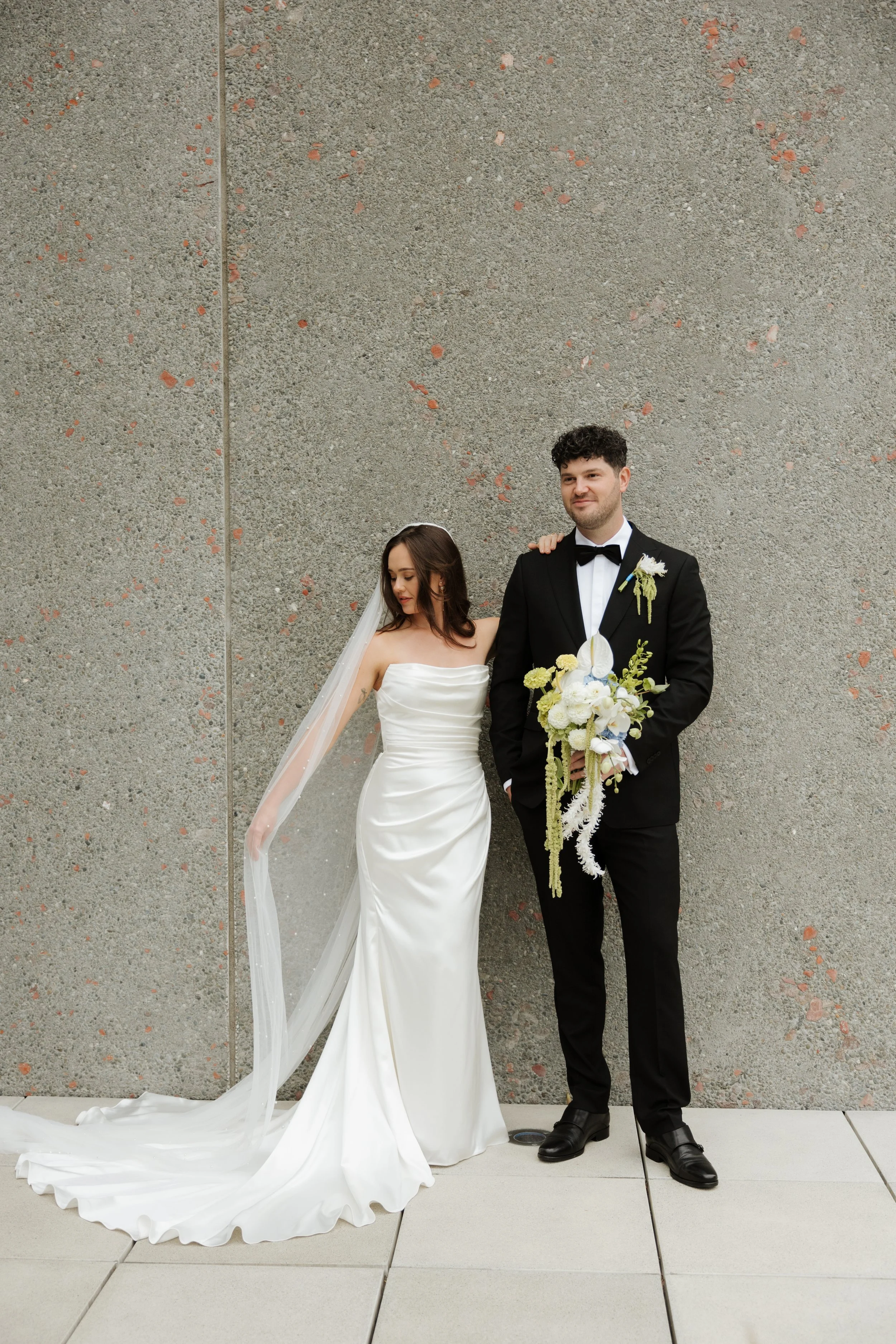 A bride and groom standing against a textured concrete wall. The bride is wearing a strapless white wedding gown with a long train and veil. The groom is dressed in a black tuxedo with a bow tie, holding a bouquet of white and yellow flowers, and has