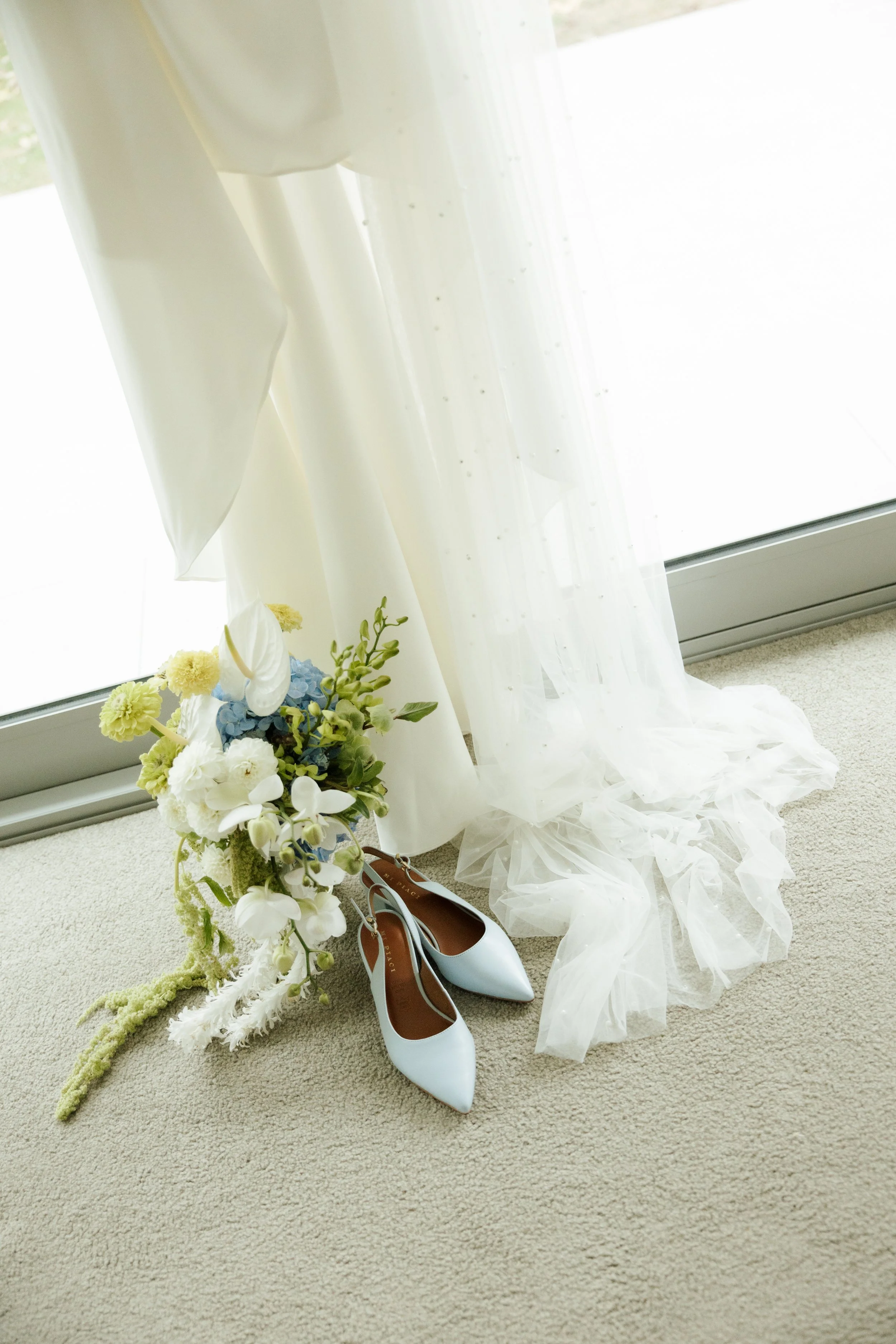 A bouquet of white and light blue flowers on the floor next to a pair of white high-heeled shoes, with sheer white curtains in the background.