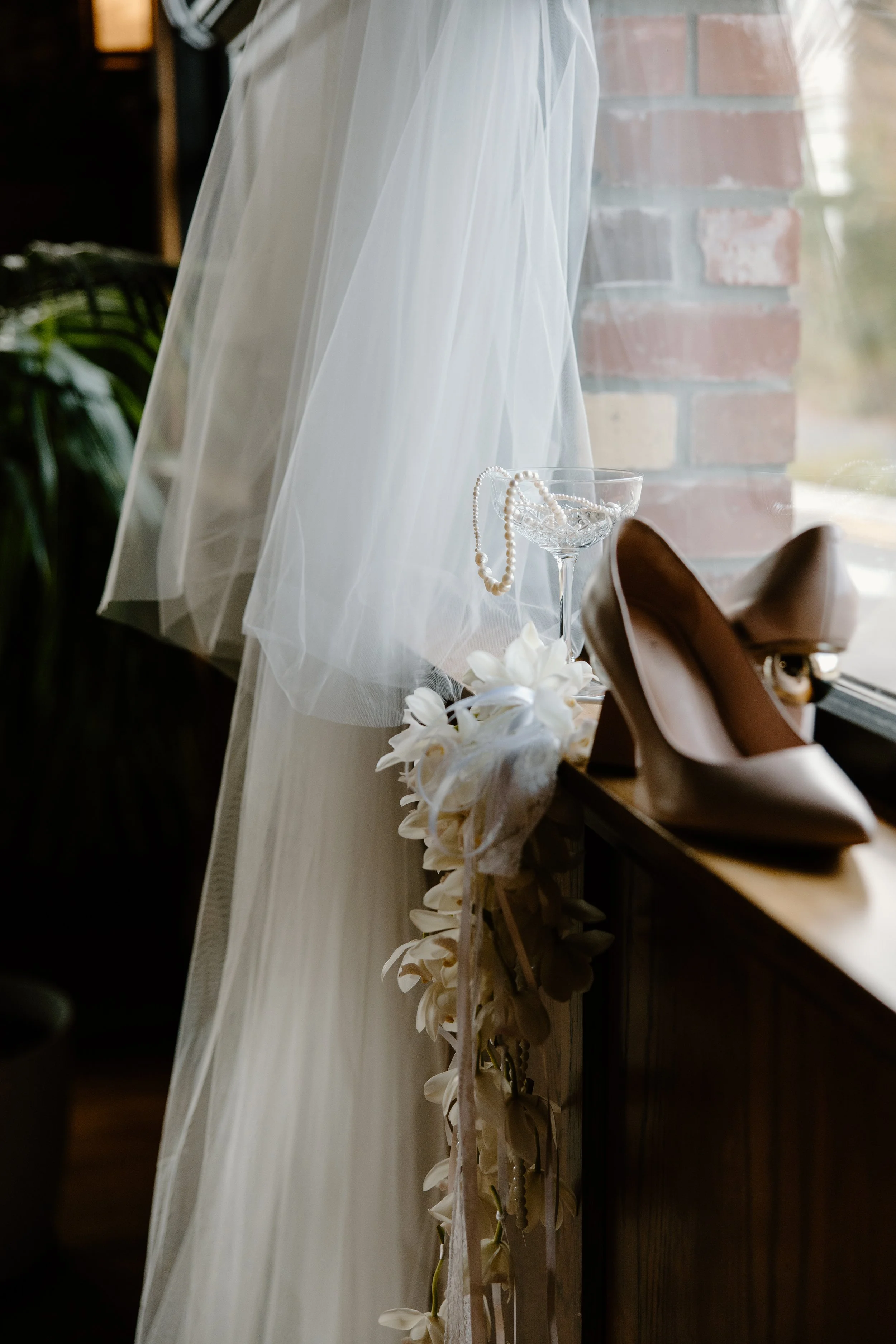 A wedding dress with veil, a string of pearls, a glass bowl, and white flowers arranged on a surface near a window with a brick wall outside.
