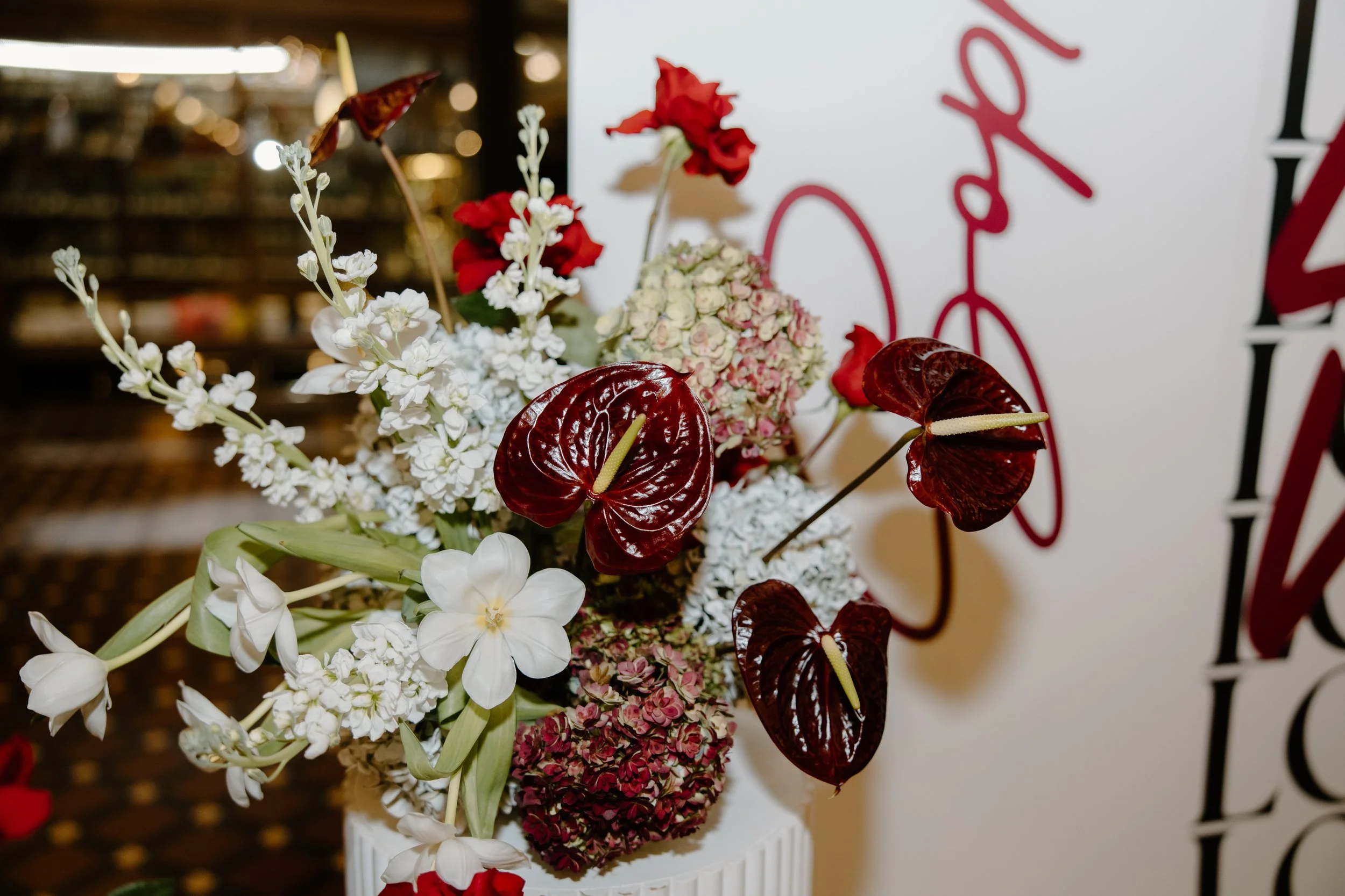 A floral arrangement featuring white, red, and pink flowers in a white vase with a blurred background that includes a wall with red and black writing.