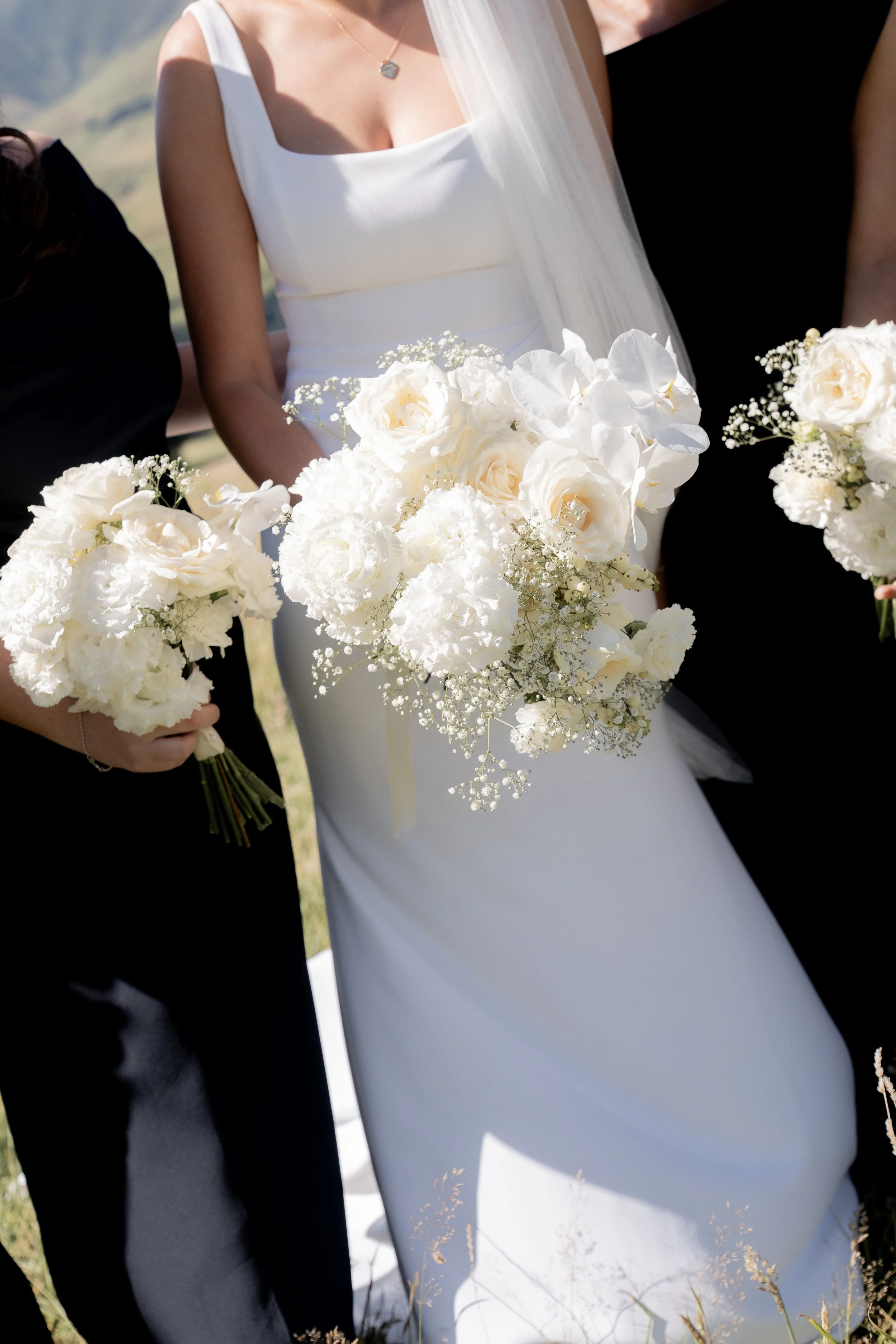 Close-up of a bride holding a bouquet of white roses, orchids, and baby's breath, standing outdoors with two people, one in a black outfit and the other in a white wedding dress, during a wedding ceremony.